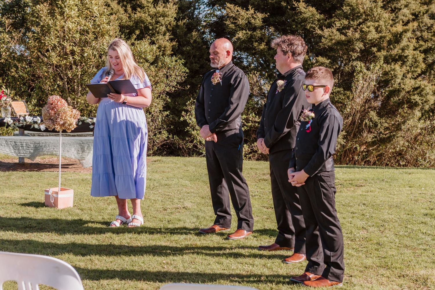A small wedding ceremony taking place on a grassy lawn with attendees in formal attire.