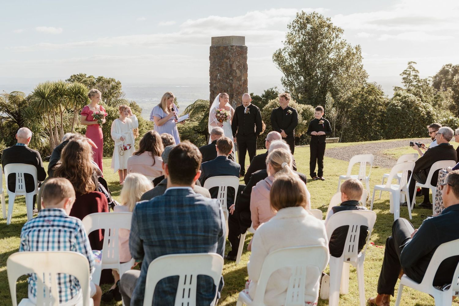 Rows of white chairs set up for an outdoor wedding ceremony with attendees gathering on a sunny afternoon.