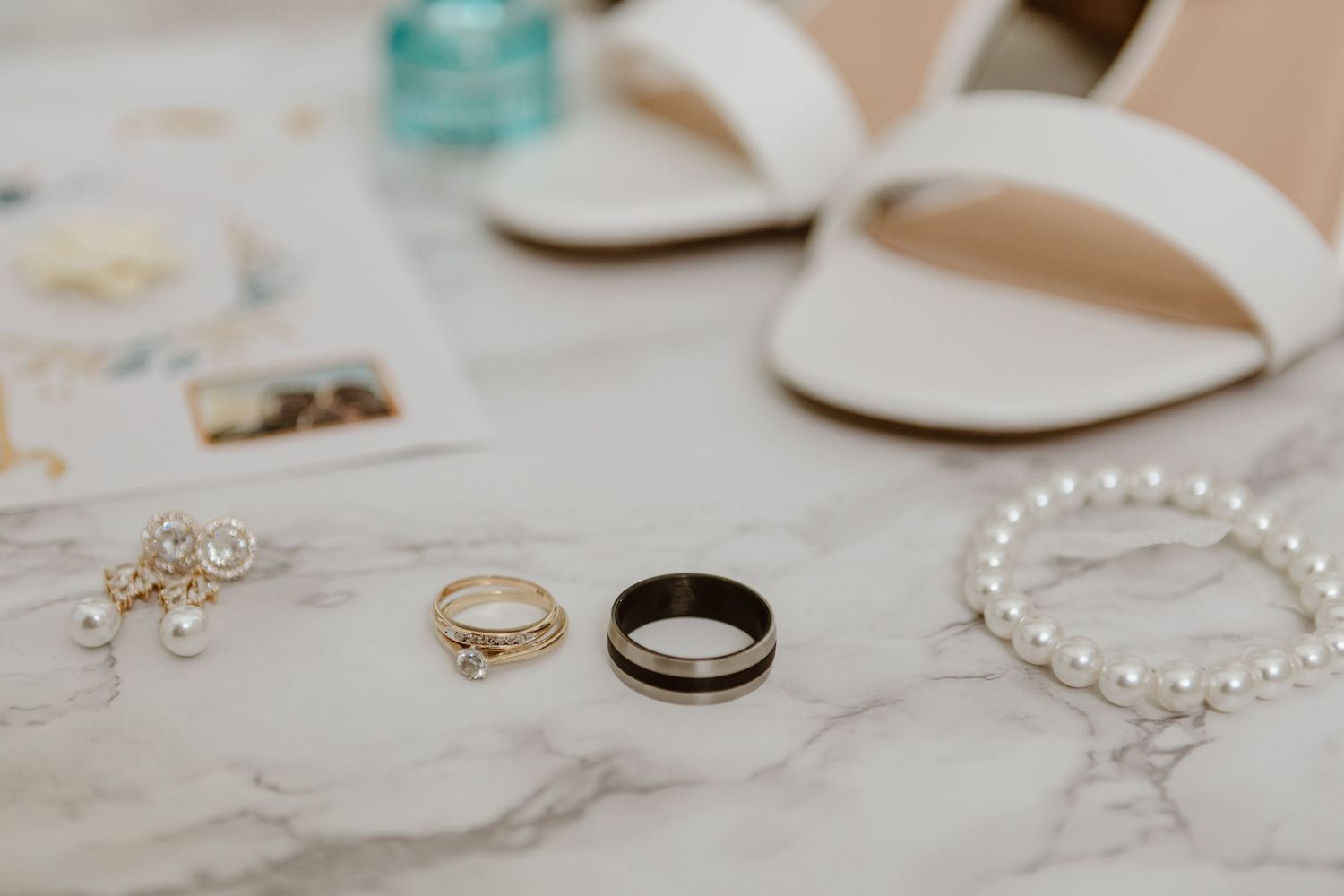 Wedding accessories including rings, pearl bracelet, and white sandals arranged on marble surface.