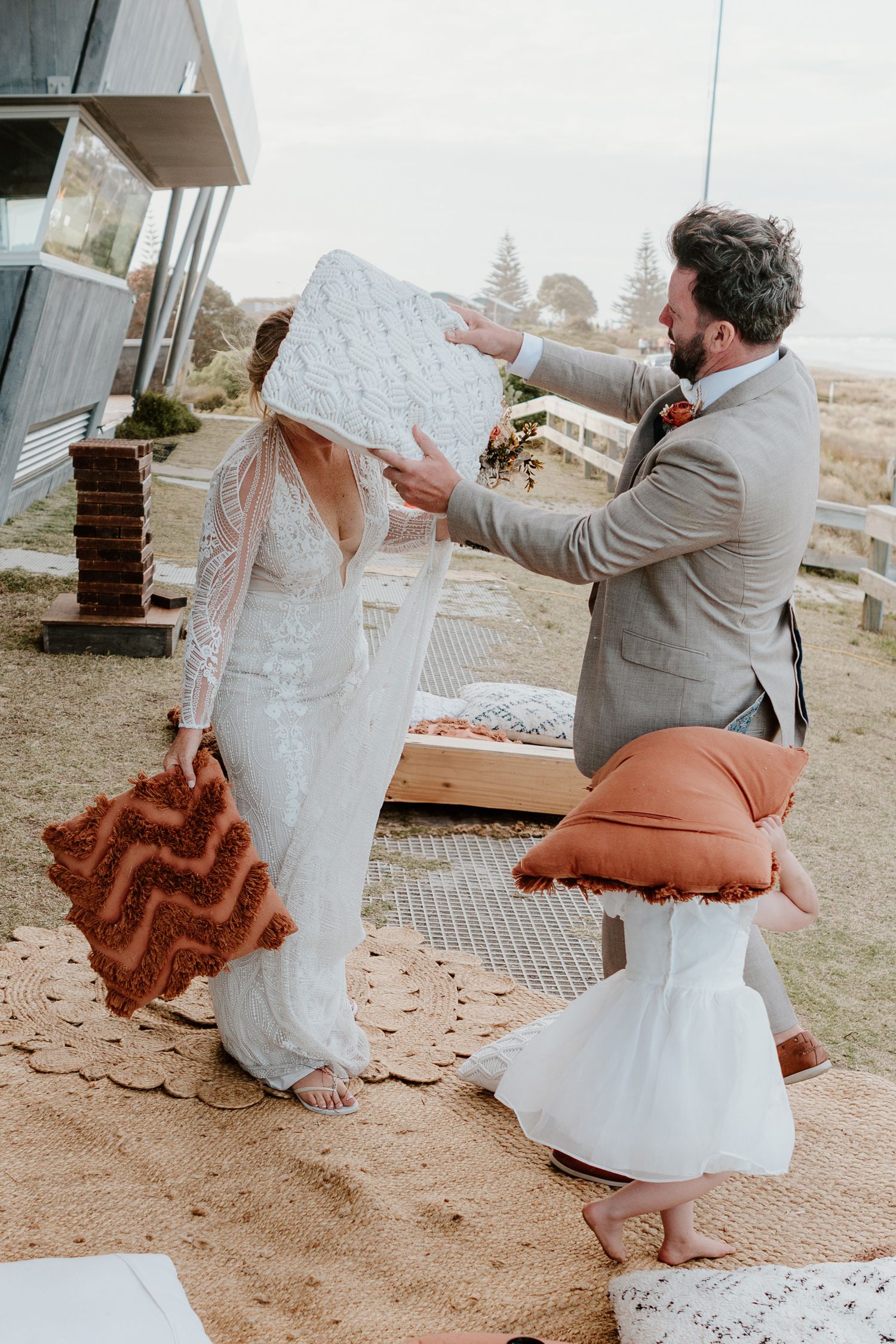 A couple having a pillow fight during an outdoor reception with boho-style cushions and decor on the beach.