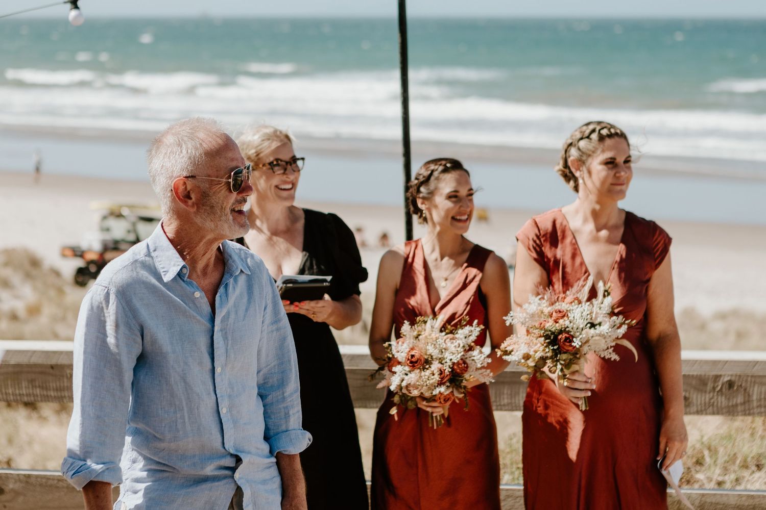 People gather on a beach near the ocean wearing casual attire with dried floral arrangements in hand.
