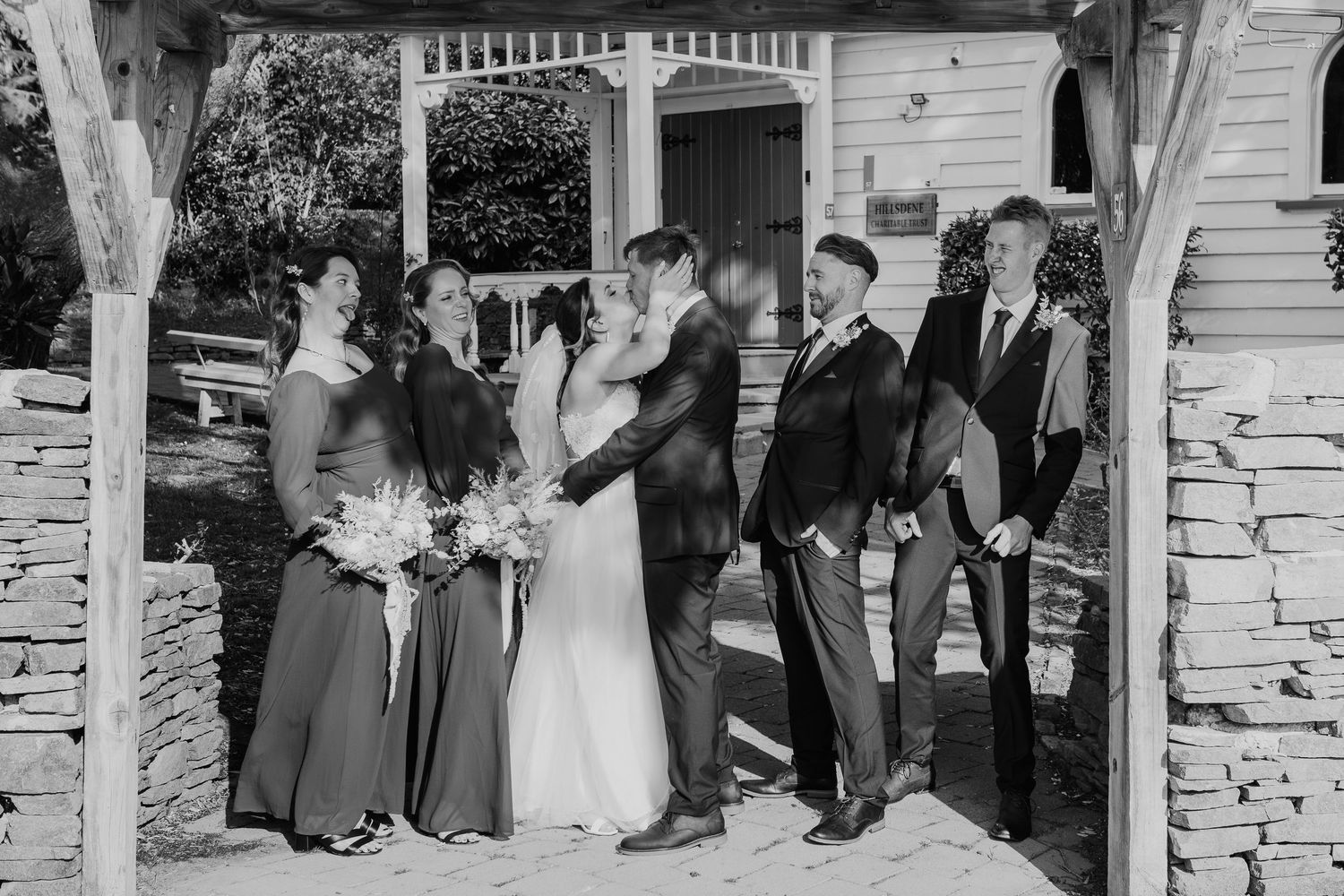 Black and white wedding party gathered under a pergola with stone pillars during an outdoor ceremony.