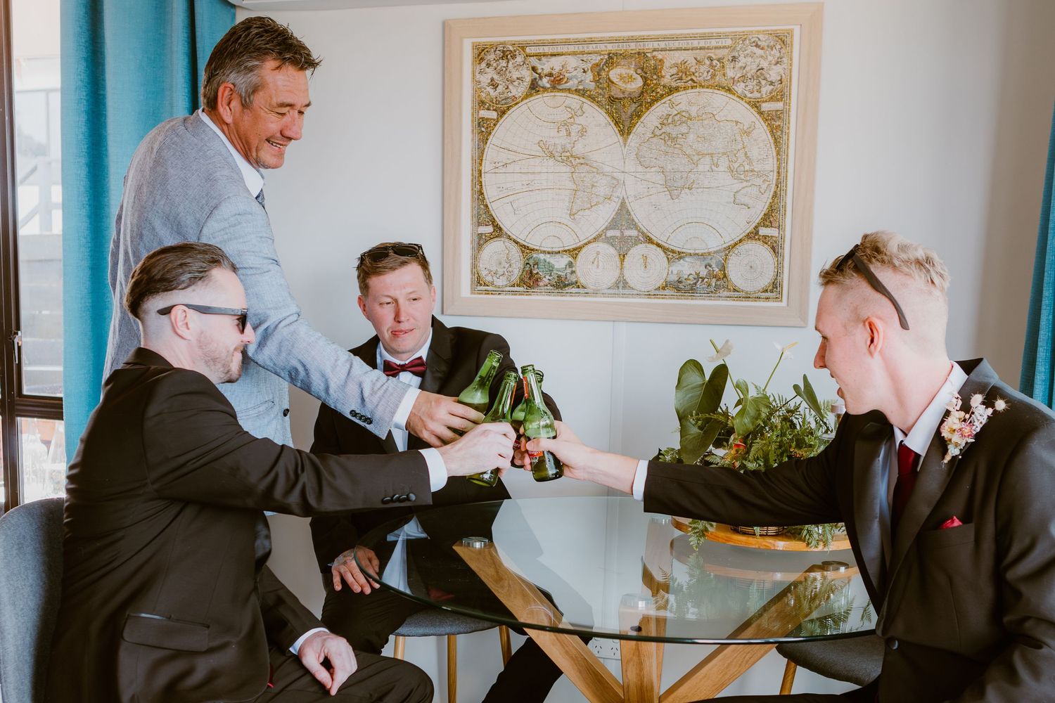 A group of people share beers around a glass table with a vintage world map on the wall behind them.