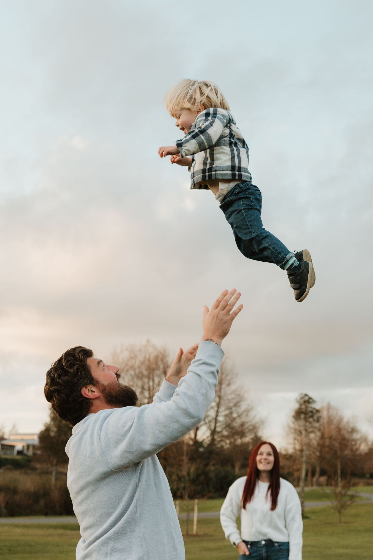 A father joyfully tosses his young blond child into the air outdoors while a smiling woman watches in the background.