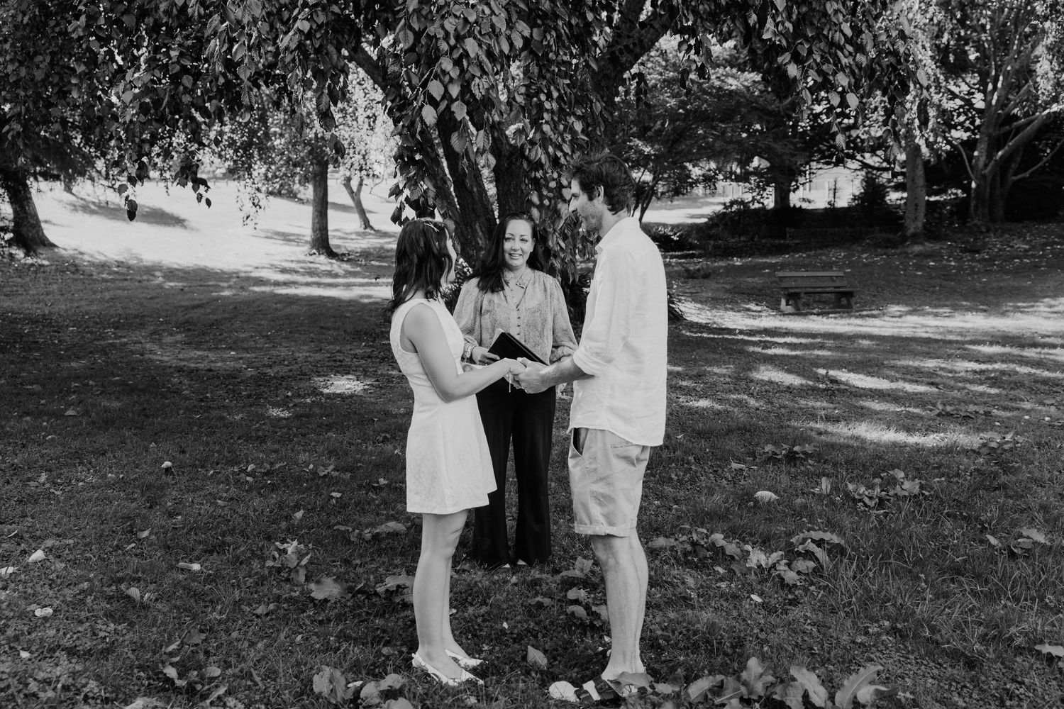 Black and white photo of three people exchanging vows outdoors under a tree during an intimate wedding ceremony.