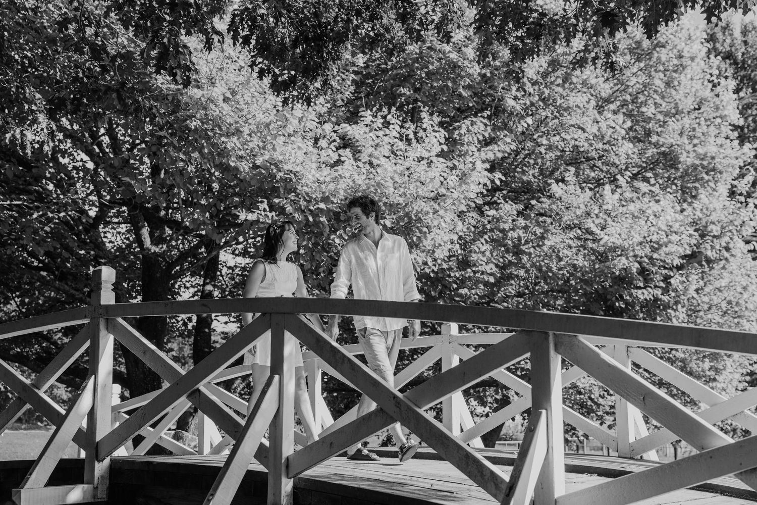 A couple stands on a wooden bridge surrounded by lush trees in a romantic black and white outdoor portrait.