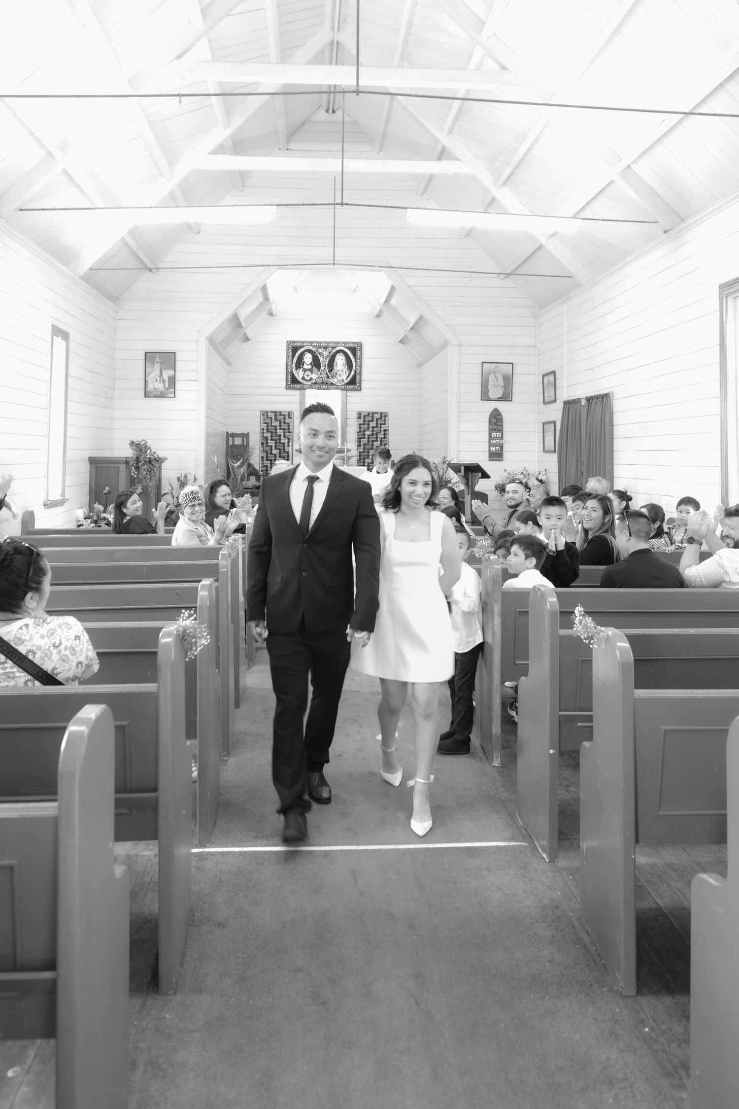A couple walks down the center aisle of a small white church with wooden pews and guests seated on either side.
