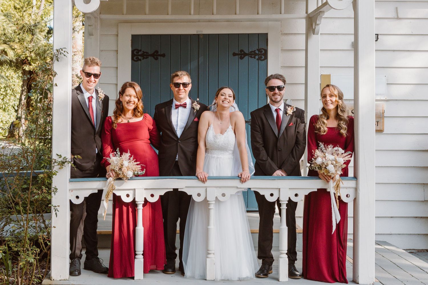 Wedding party in formal attire poses on white porch with bridesmaids in burgundy dresses and groomsmen in black suits.