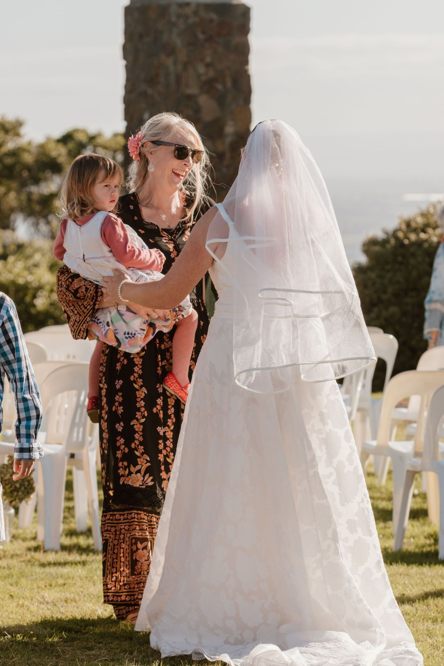 People gather at an outdoor wedding ceremony with white chairs and a bride in a flowing white gown with veil.
