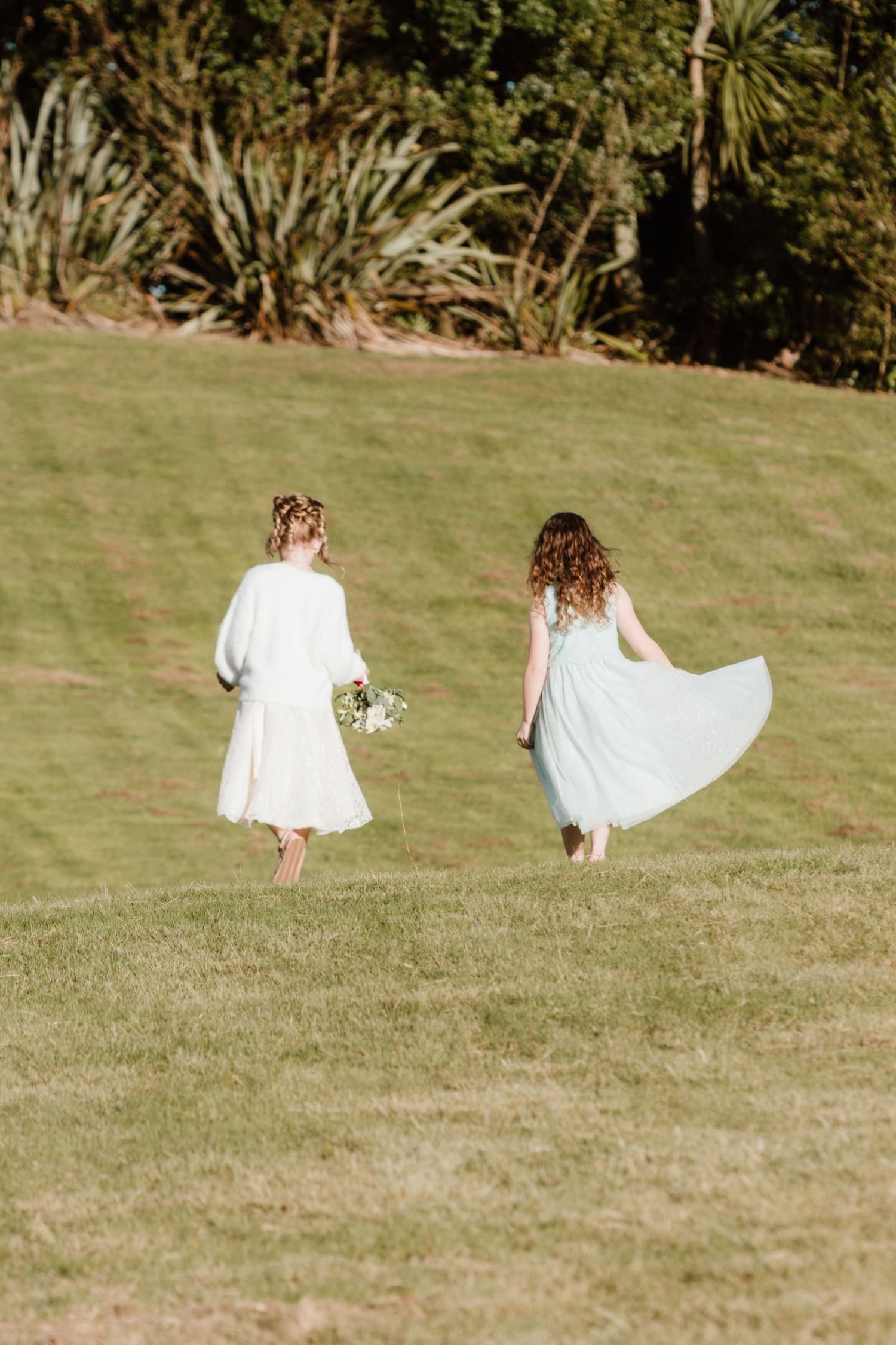 Two figures in white dresses spin and twirl playfully on a sunny grassy hillside during a photoshoot.