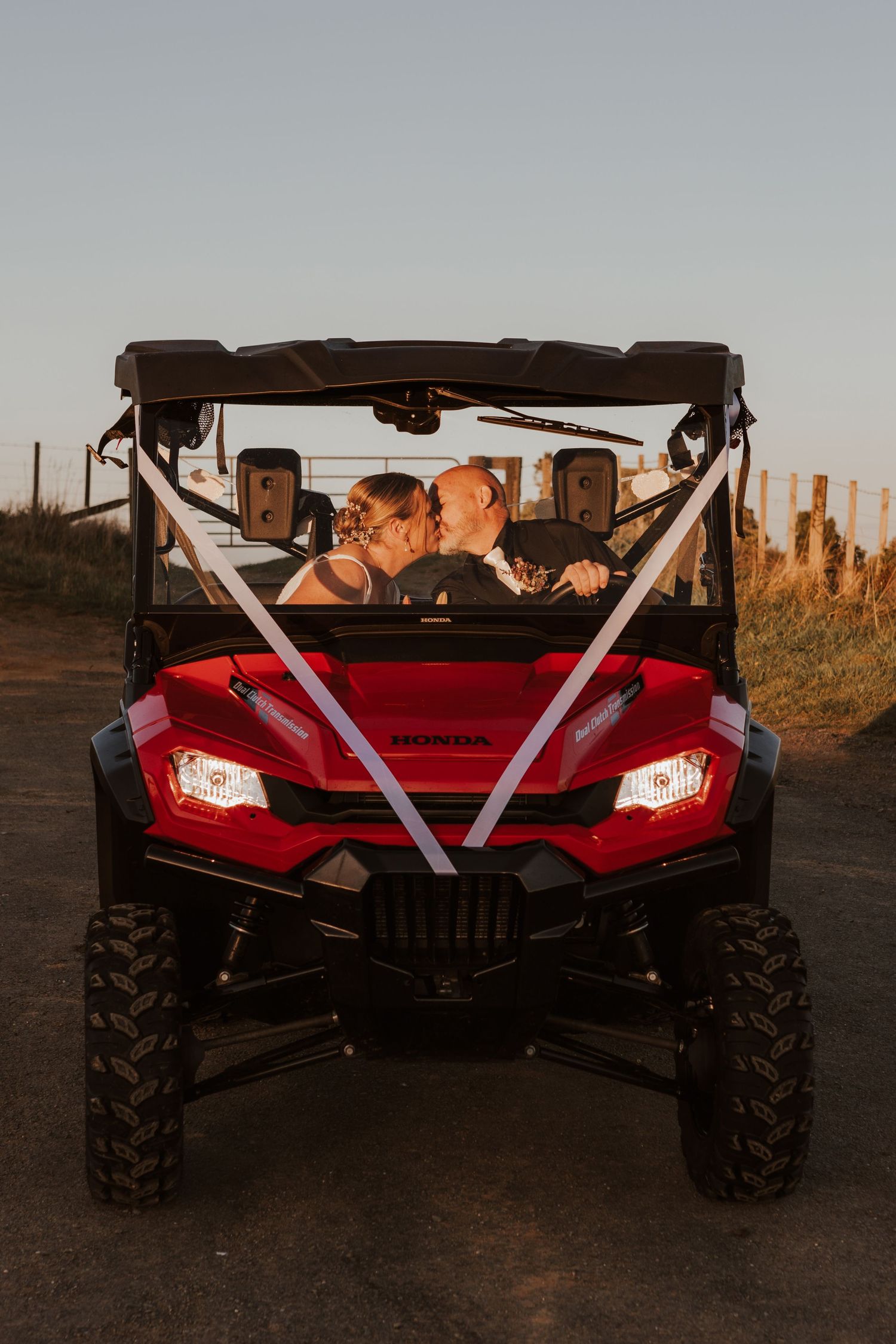 A red off-road vehicle decorated with white ribbons is parked at sunset with headlights illuminated.