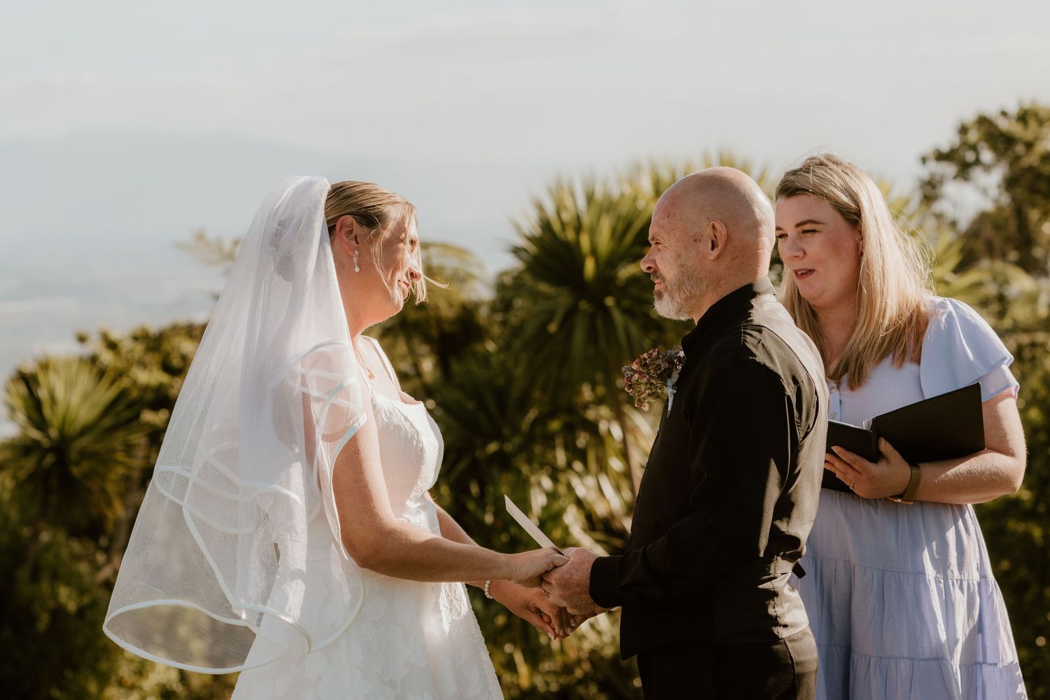 A couple participates in a wedding ceremony outdoors with palm trees in the background during golden hour.
