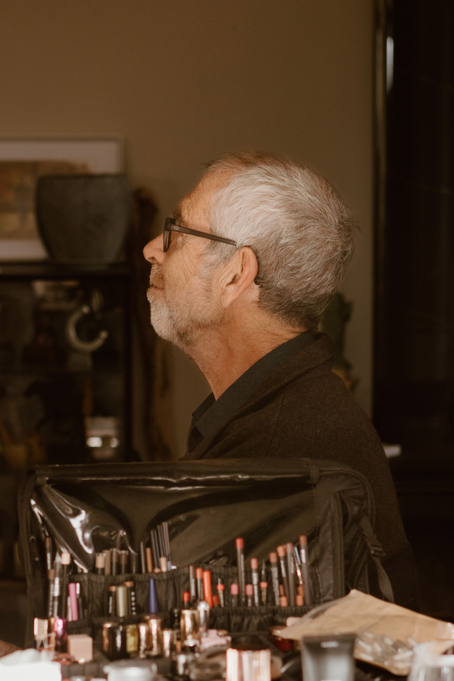 Side profile view of someone with white hair and glasses near a makeup station with lipsticks and beauty products.