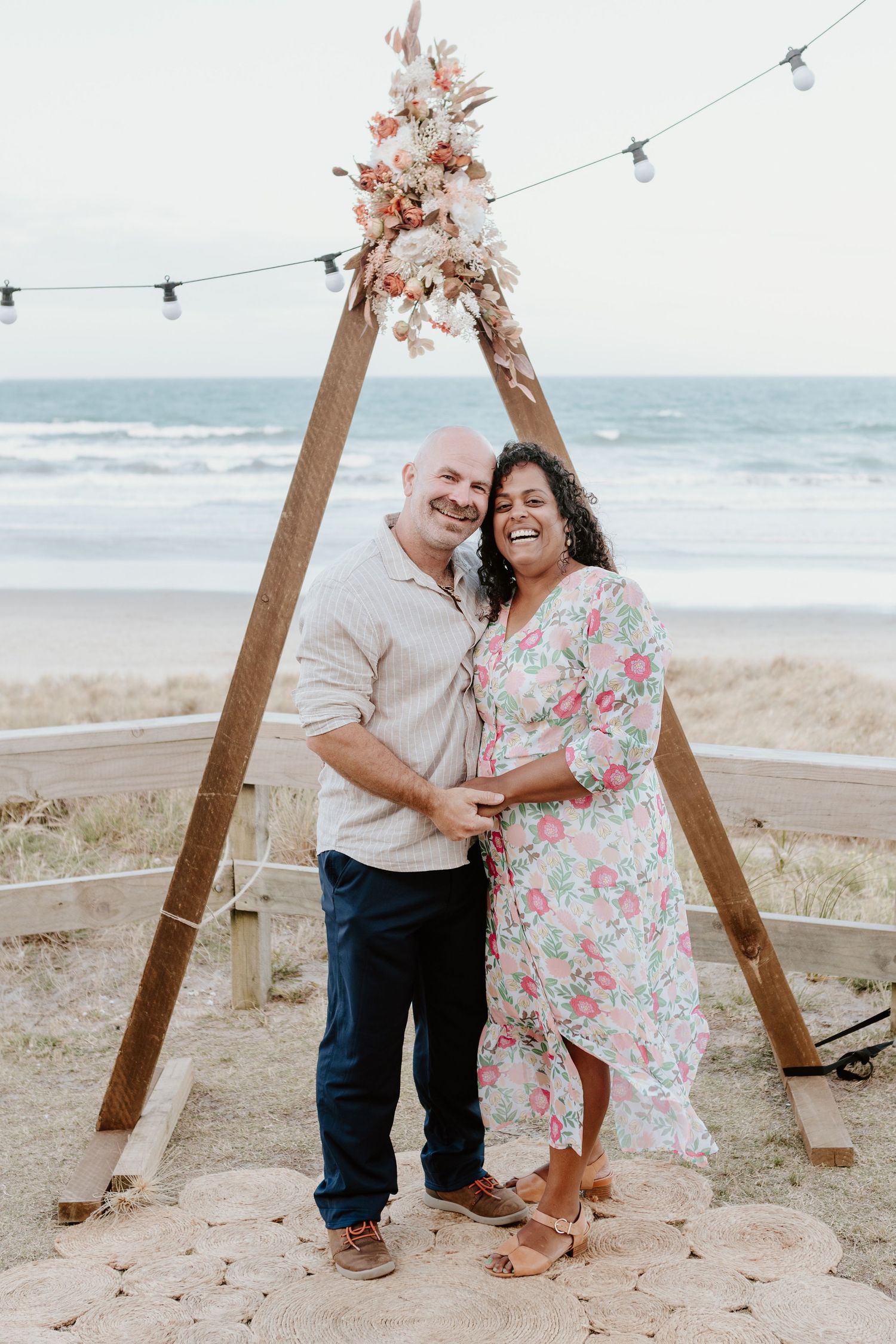 A couple poses under a wooden arch with floral decor and string lights on a beach at sunset.