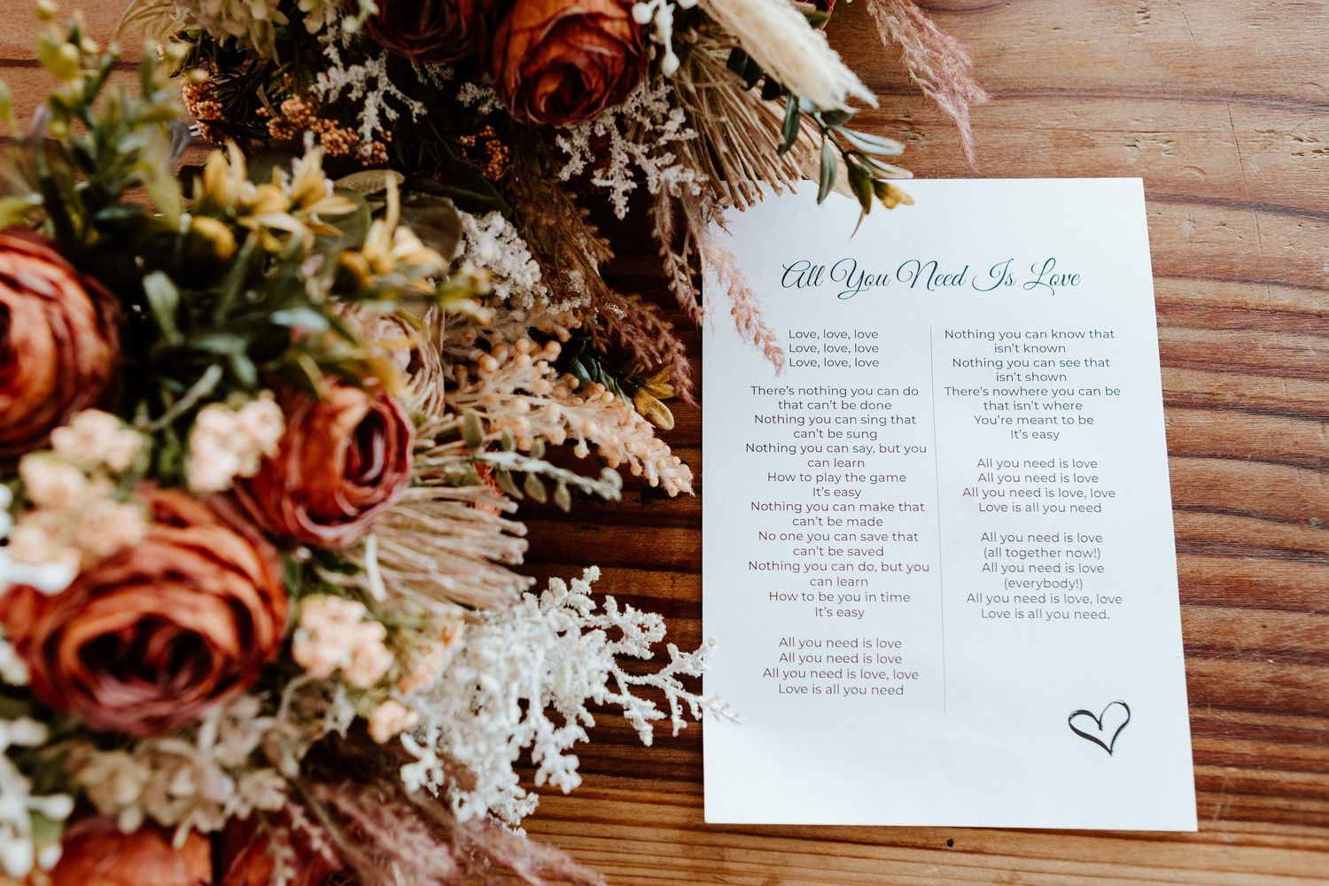 Wedding program laid on wooden surface beside autumn-colored floral arrangement with roses and white flowers.
