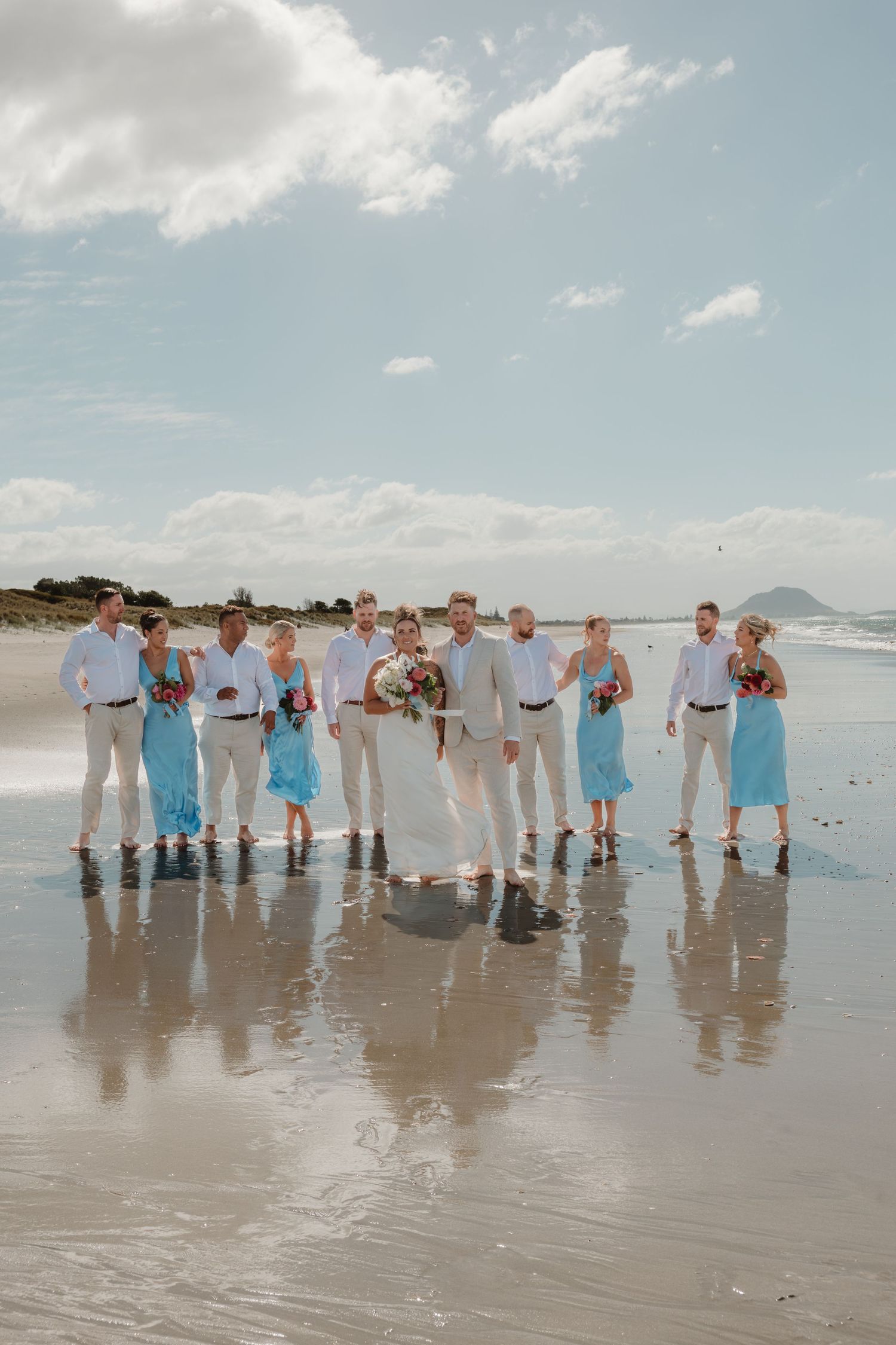 Wedding party in blue dresses and beige suits posing on a sunny beach with wet sand reflections and a mountain backdrop.