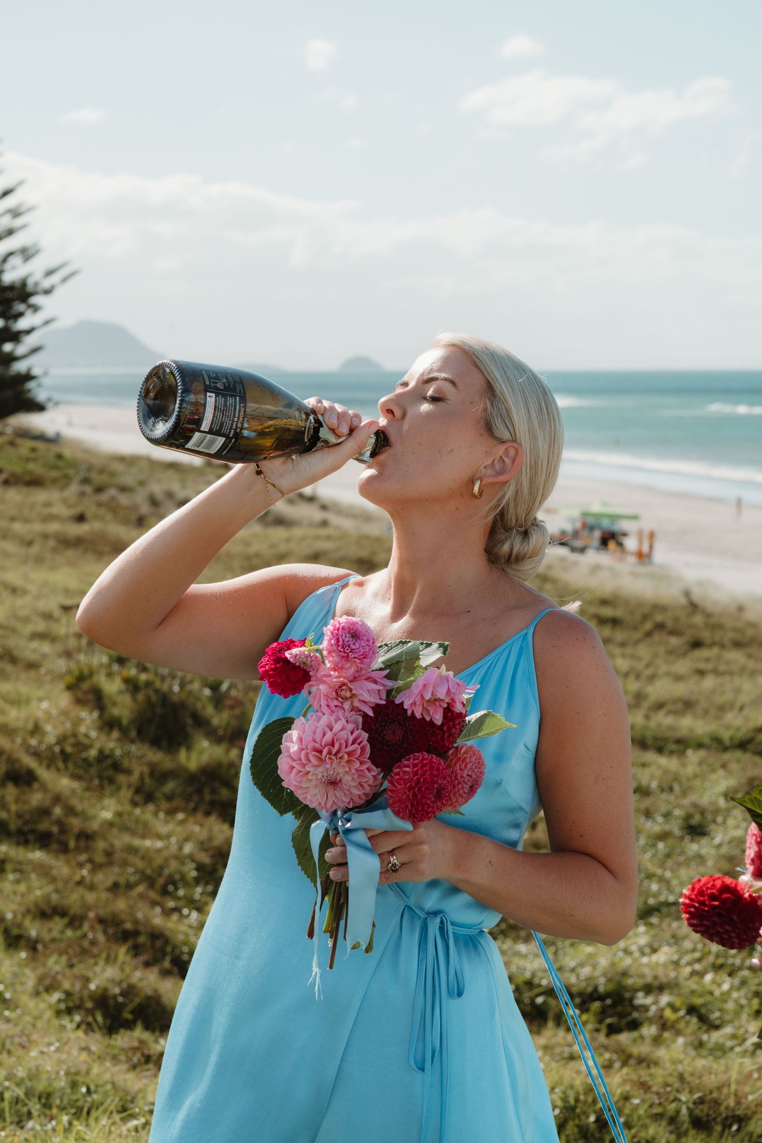 Blonde woman in blue dress drinking champagne bottle while holding pink floral bouquet on grassy beachside hill.