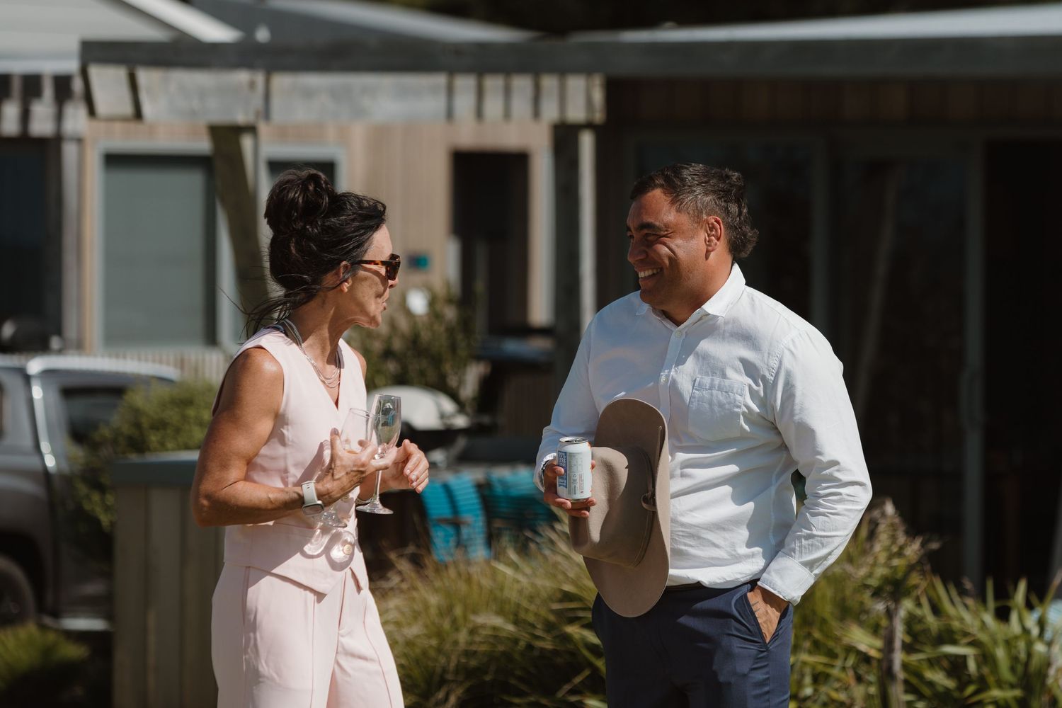A man and woman in smart casual attire chat outdoors holding drinks near a modern building.