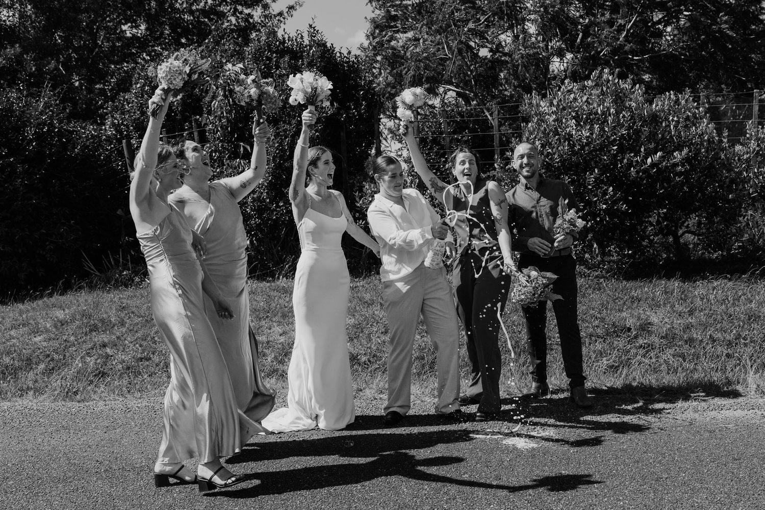 Black and white photo of a bride and wedding party joyfully raising bouquets outdoors on a sunny day.