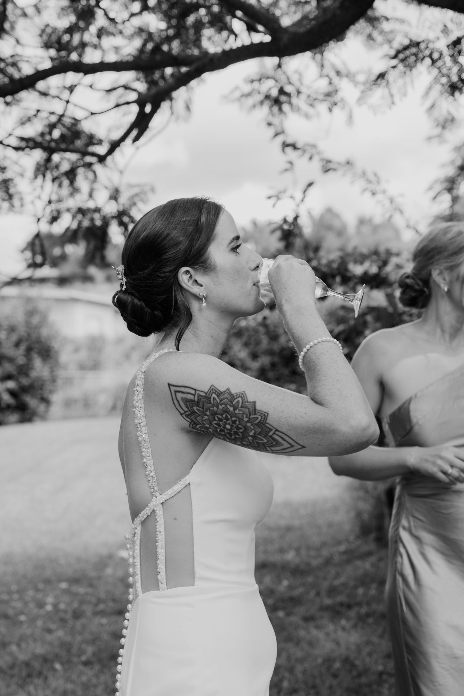 Bride with mandala arm tattoo sipping champagne in white wedding gown outdoors in black and white photo.