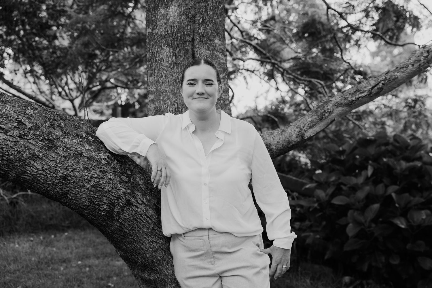 Woman smiling and leaning against a tree outdoors in a black and white portrait photo.