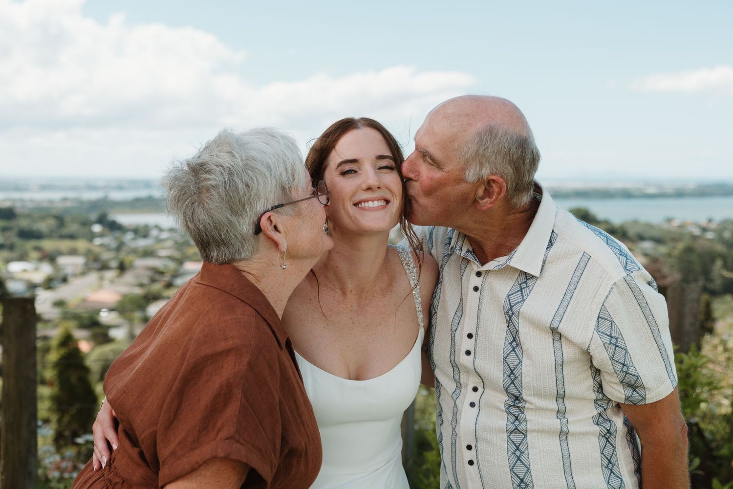Elderly couple kissing young bride on cheeks outdoors with scenic landscape view in background.