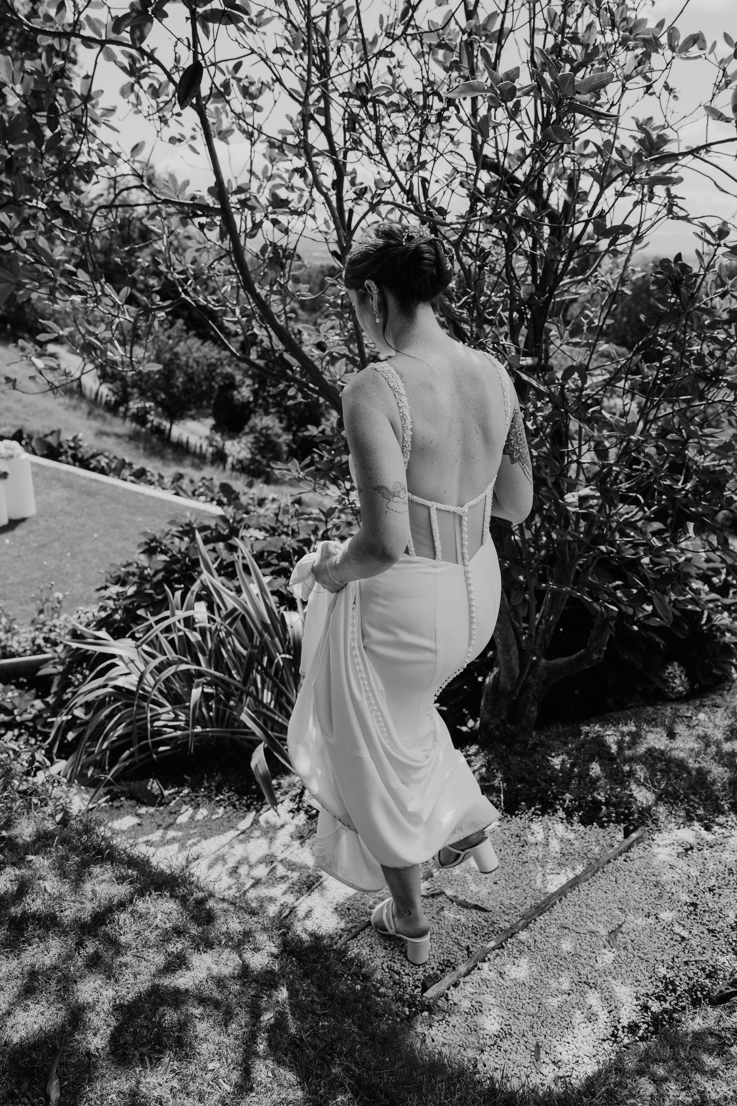 Black and white photo of a bride in a backless white lace wedding gown walking through a lush garden.
