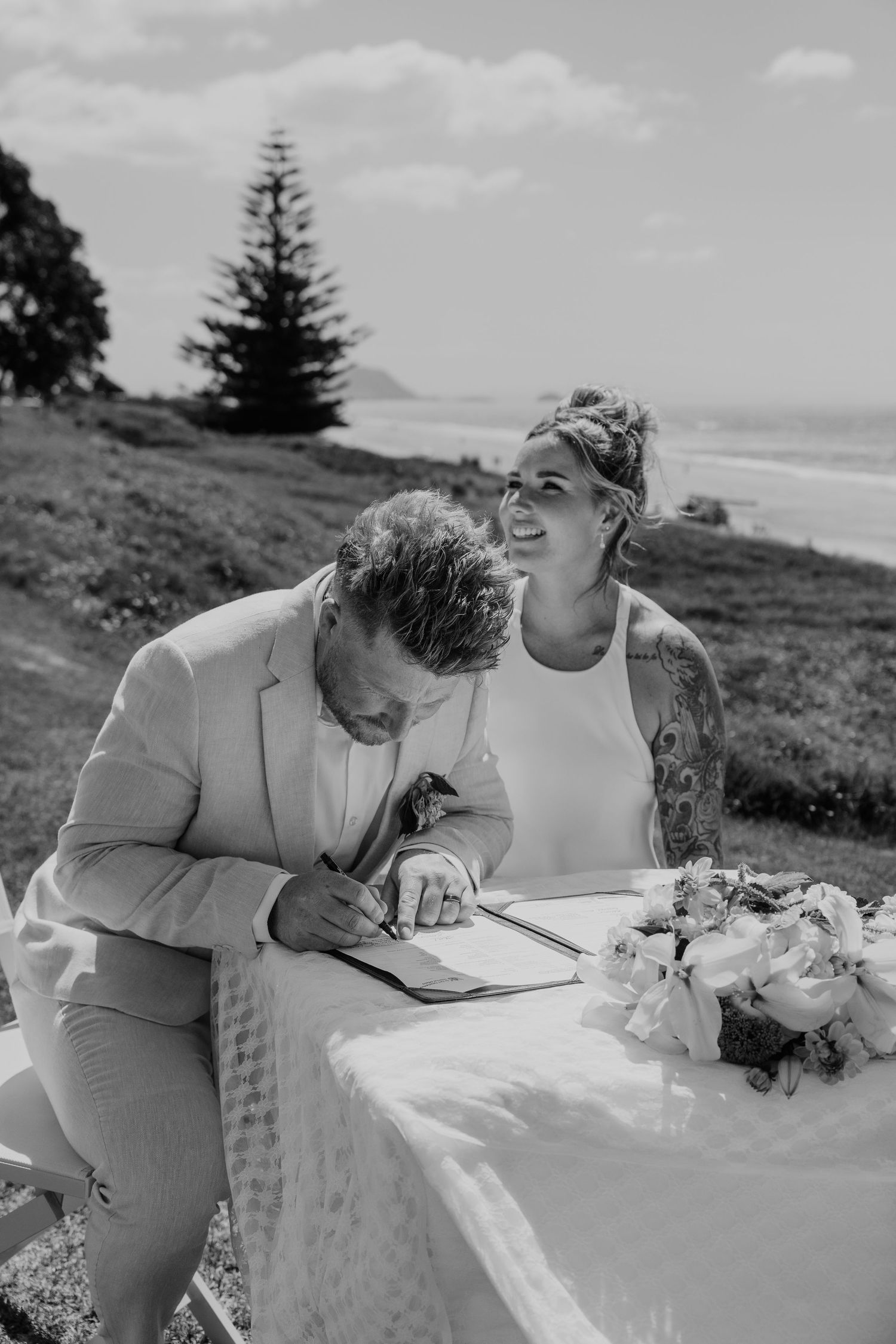 Black and white photo of a groom signing wedding registry while his smiling bride watches at an outdoor coastal ceremony.