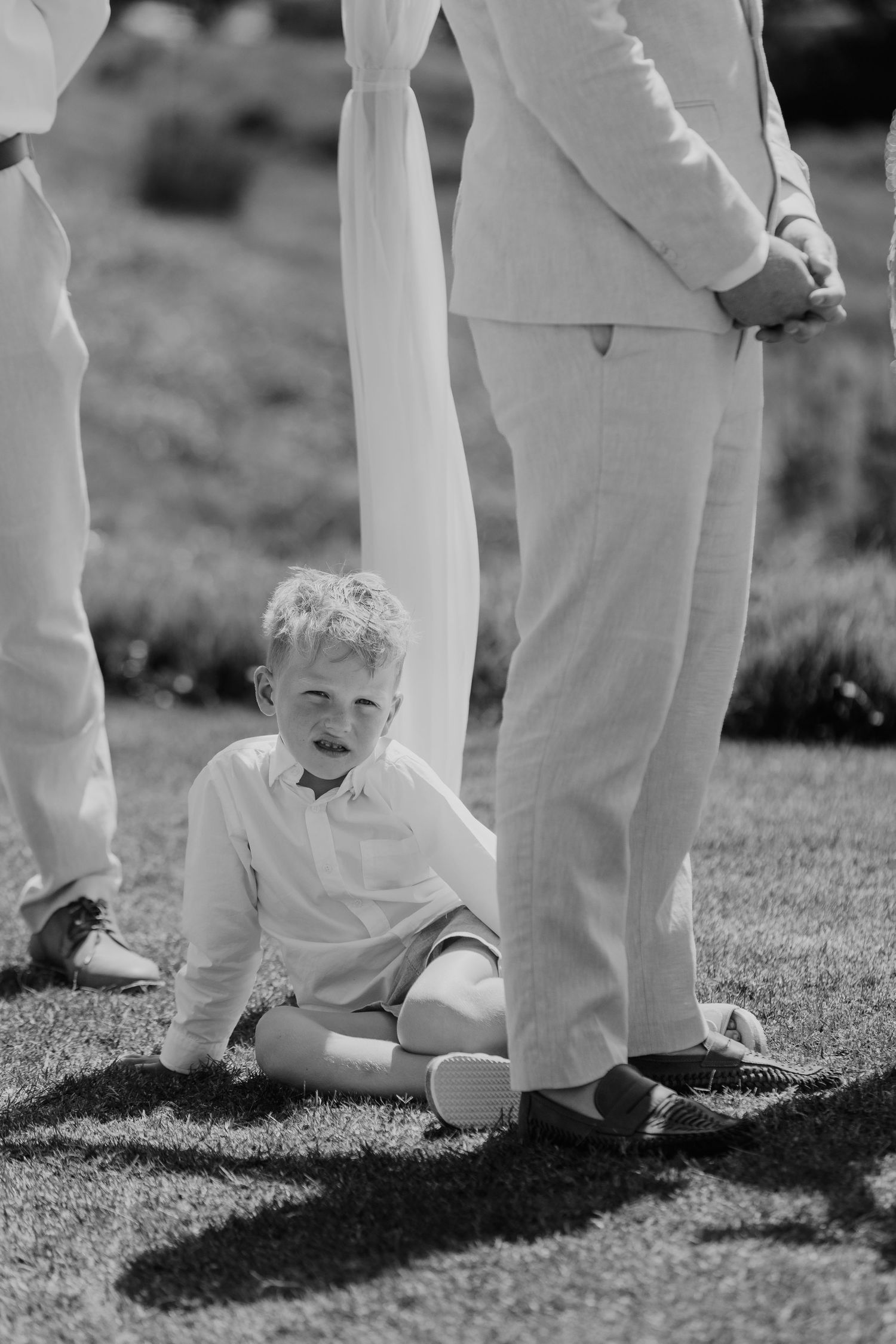 Black and white photo of a young boy sitting on grass between adults in formal wear at an outdoor wedding ceremony.