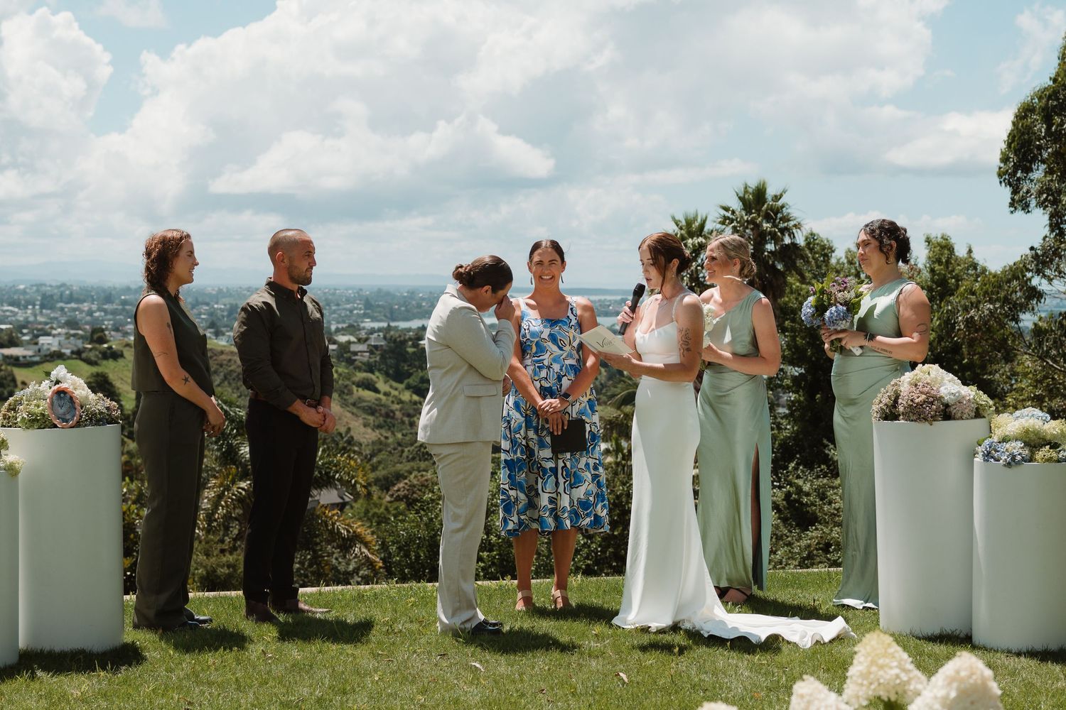 Outdoor wedding ceremony on a hilltop with bride, groom, officiant, and wedding party against a scenic city backdrop.