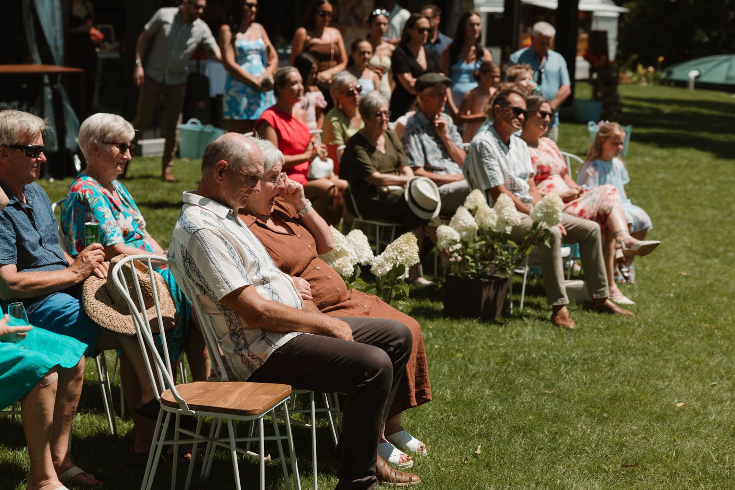 Guests seated on chairs outdoors at a sunny garden ceremony, with lush green trees in the background.