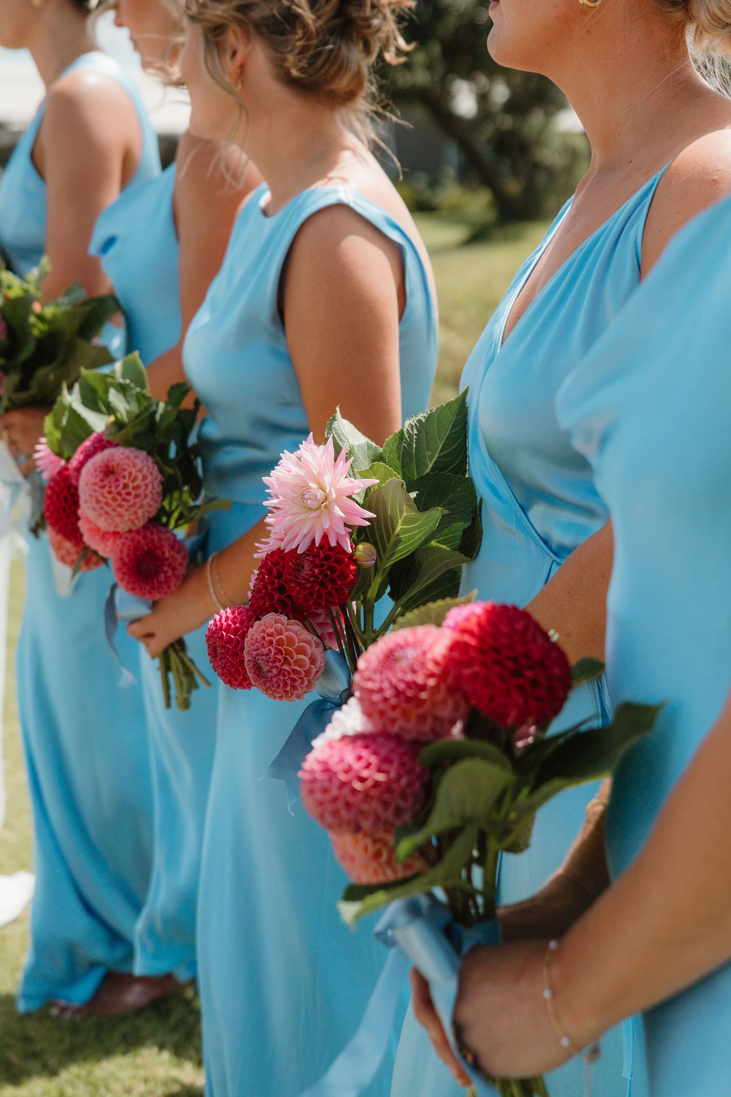 Bridesmaids in light blue gowns holding vibrant pink and red dahlia bouquets at an outdoor wedding ceremony.