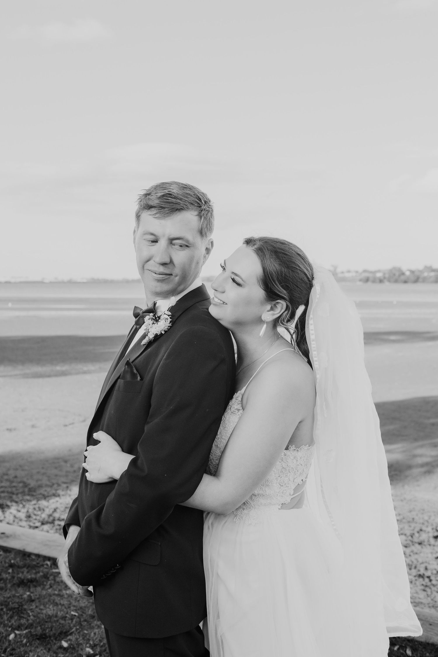 Intimate black and white portrait of newlyweds embracing against a waterfront backdrop.