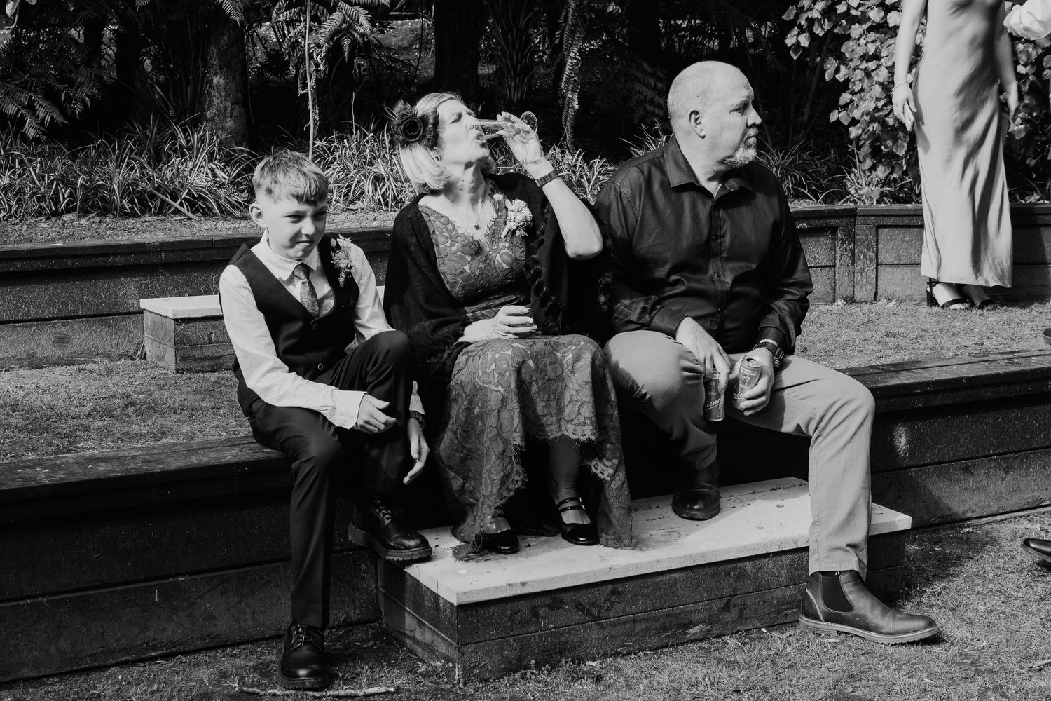 Three guests sit casually on wooden steps during an outdoor wedding.