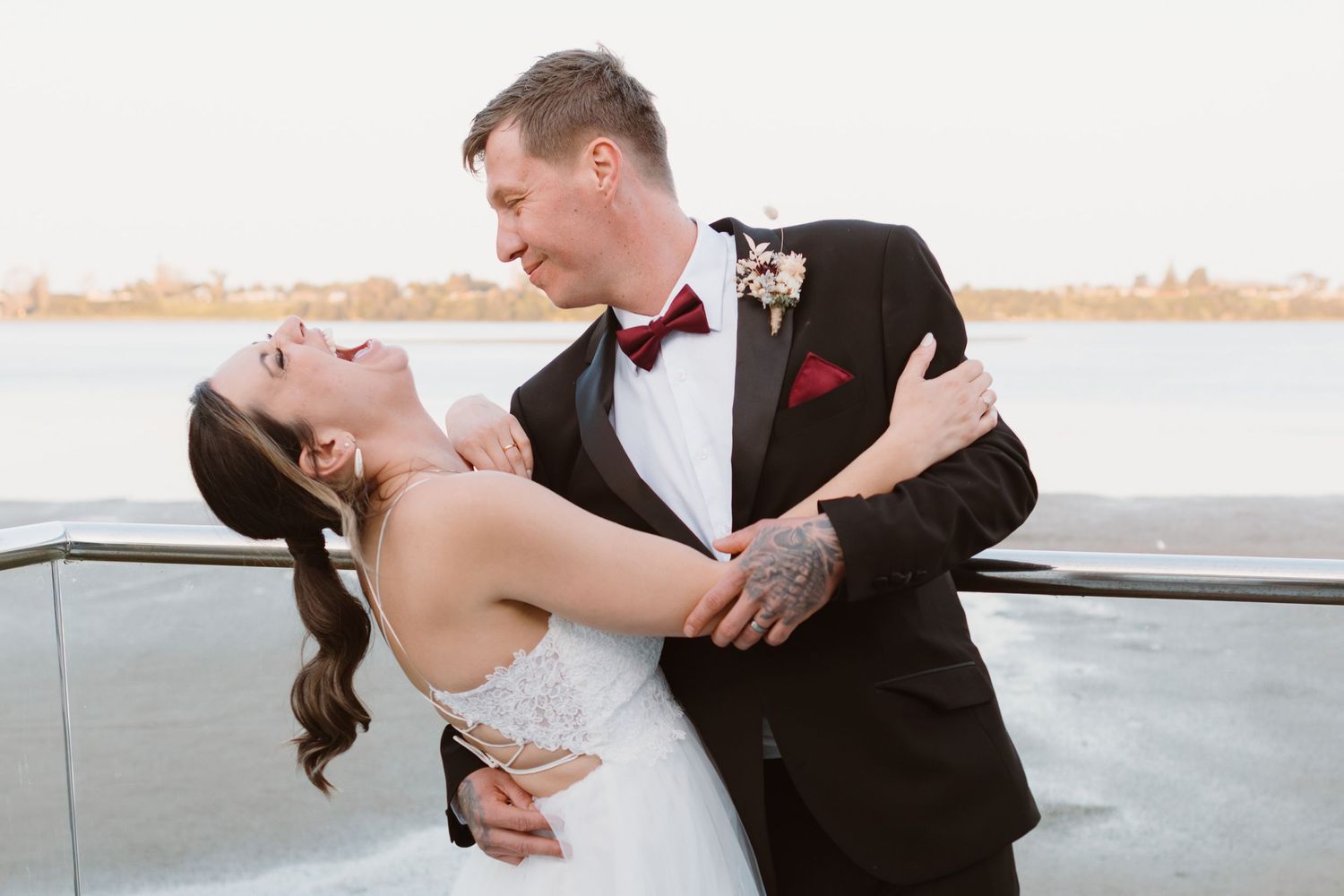 Elegant couple in formal wedding attire share a romantic dip pose at sunset by the water. Tauranga  wedding, The Waters Edge Venue