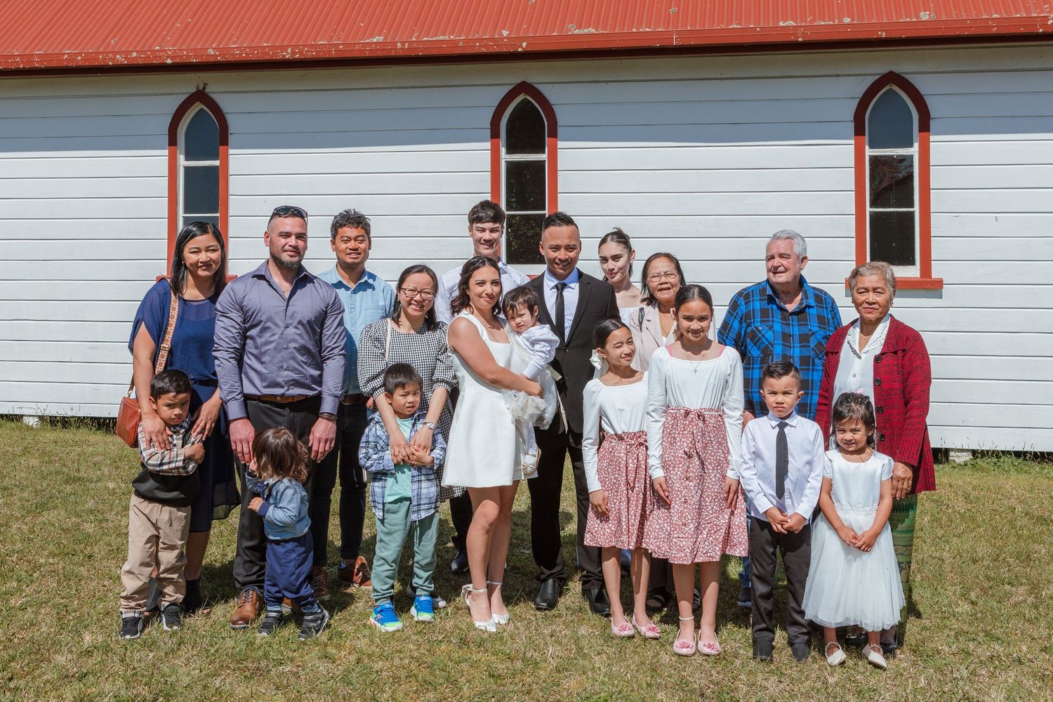 Large group photo of wedding attendees in front of a white church with red roof.