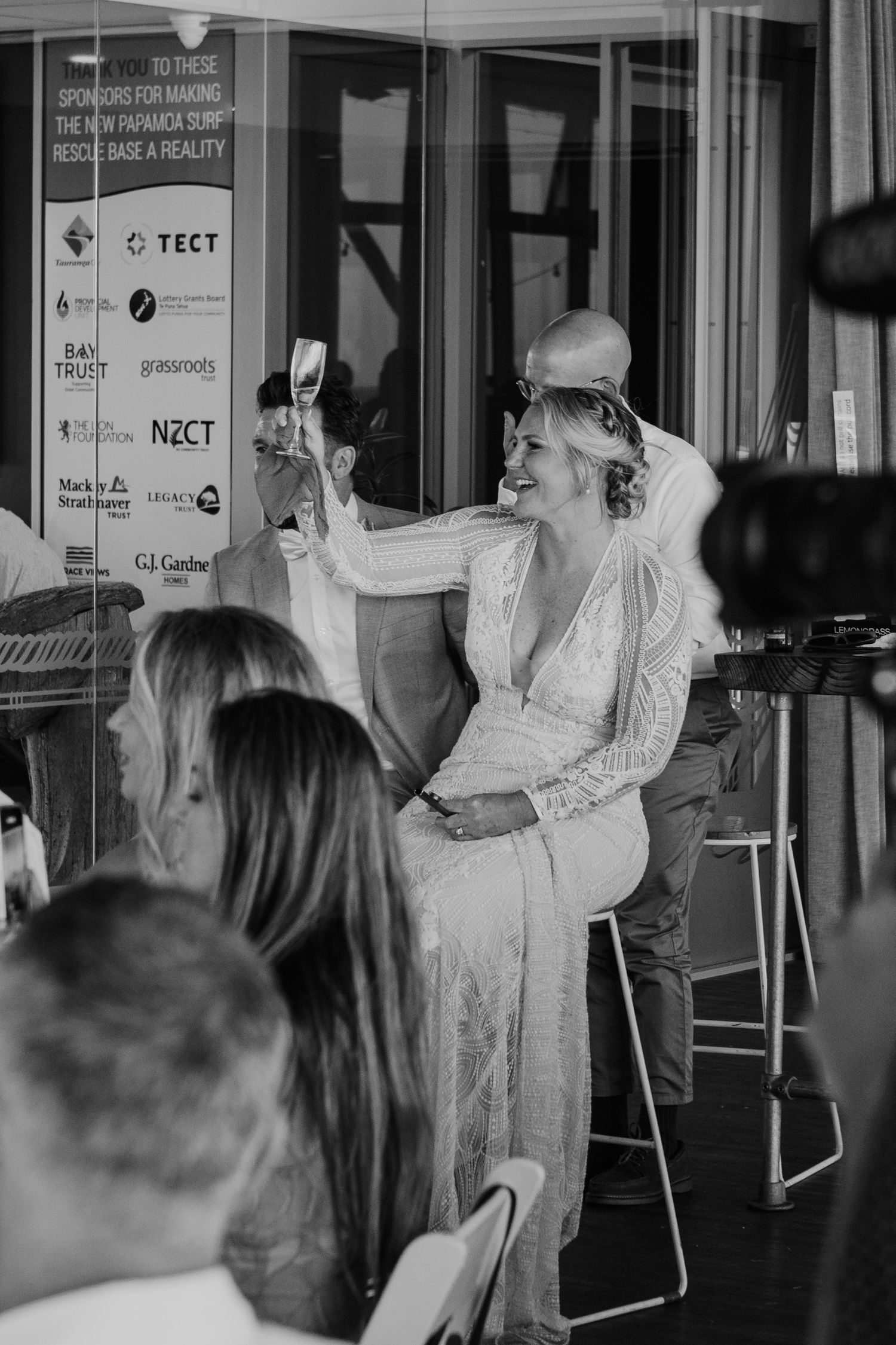 A bride in a long-sleeved lace dress stands and gestures during her wedding speech. Papamoa Surf Lifesaving Club Wedding