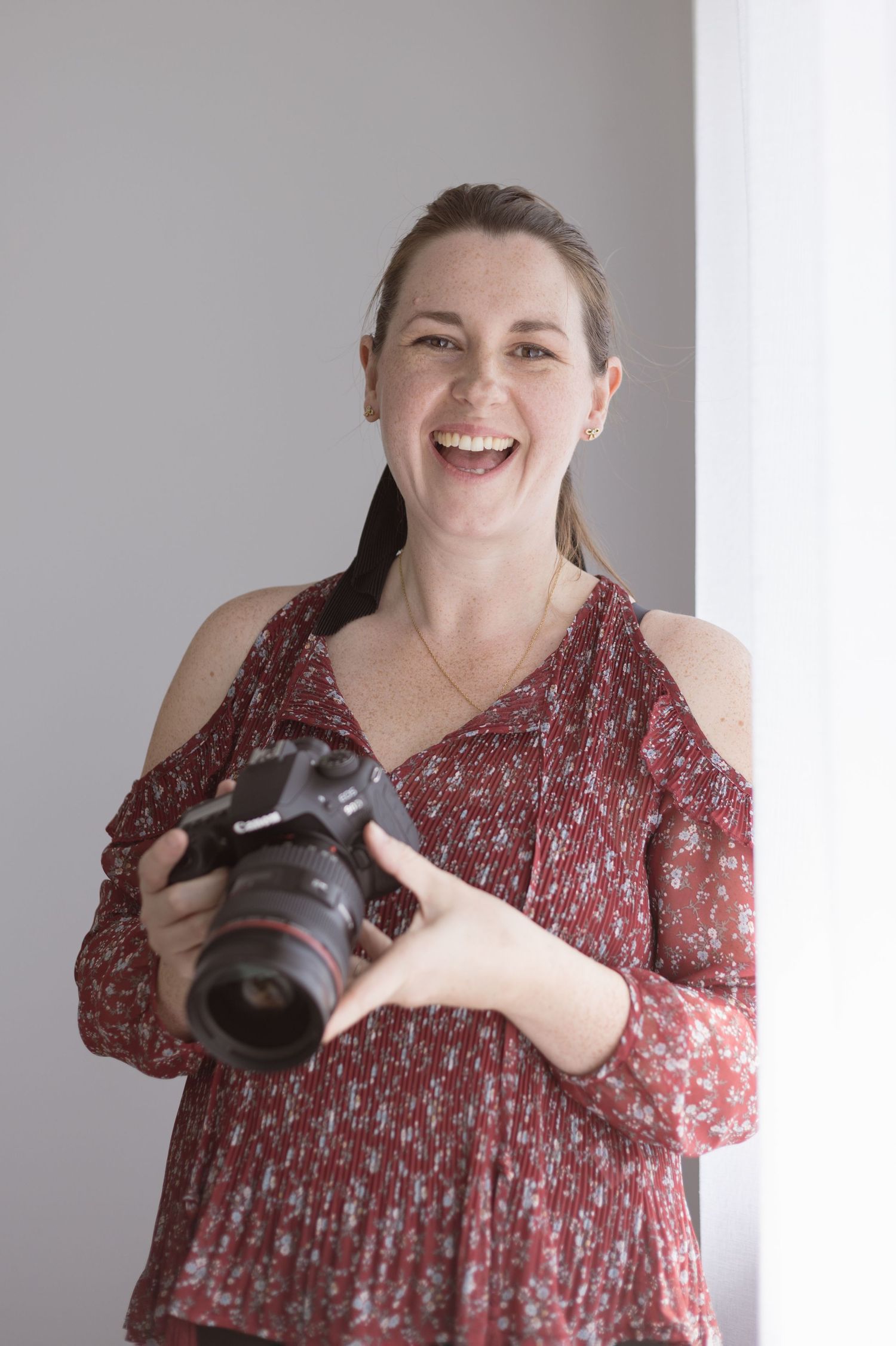 Portrait of a photographer wearing a red floral cold-shoulder top while holding a DSLR camera and smiling. Tauranga photographer