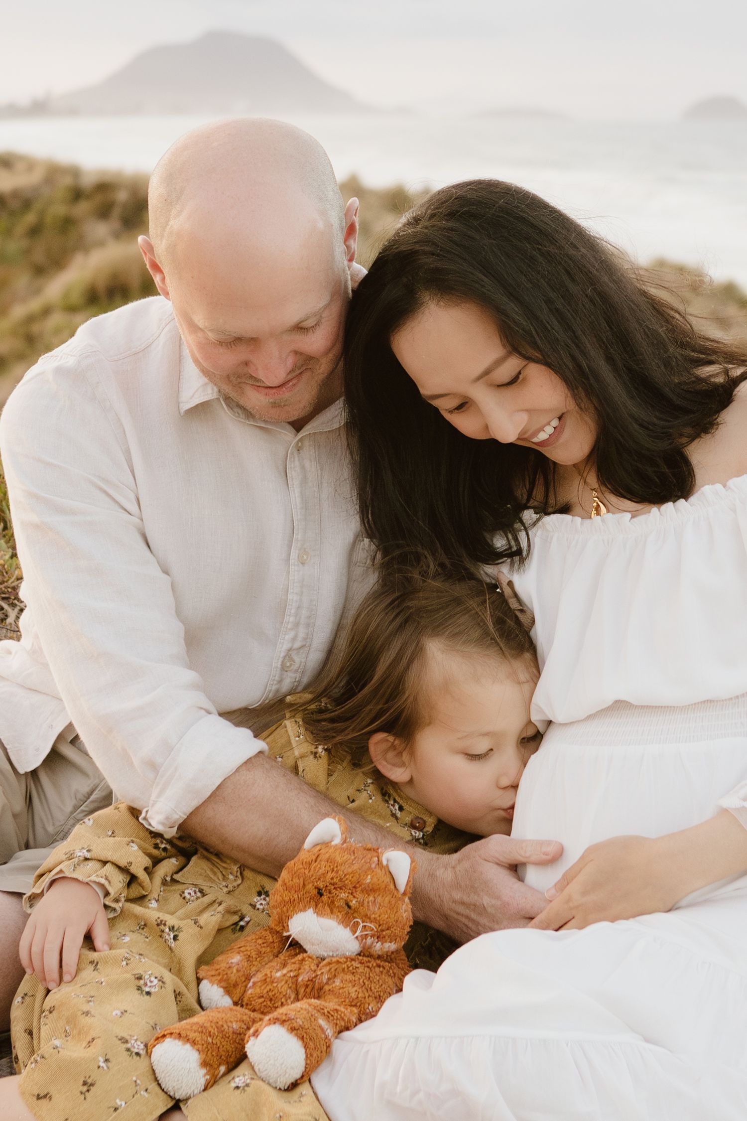 A family sits together on a hillside at sunset with a teddy bear, sharing an intimate maternity moment. Tauranga family photographer Papamoa