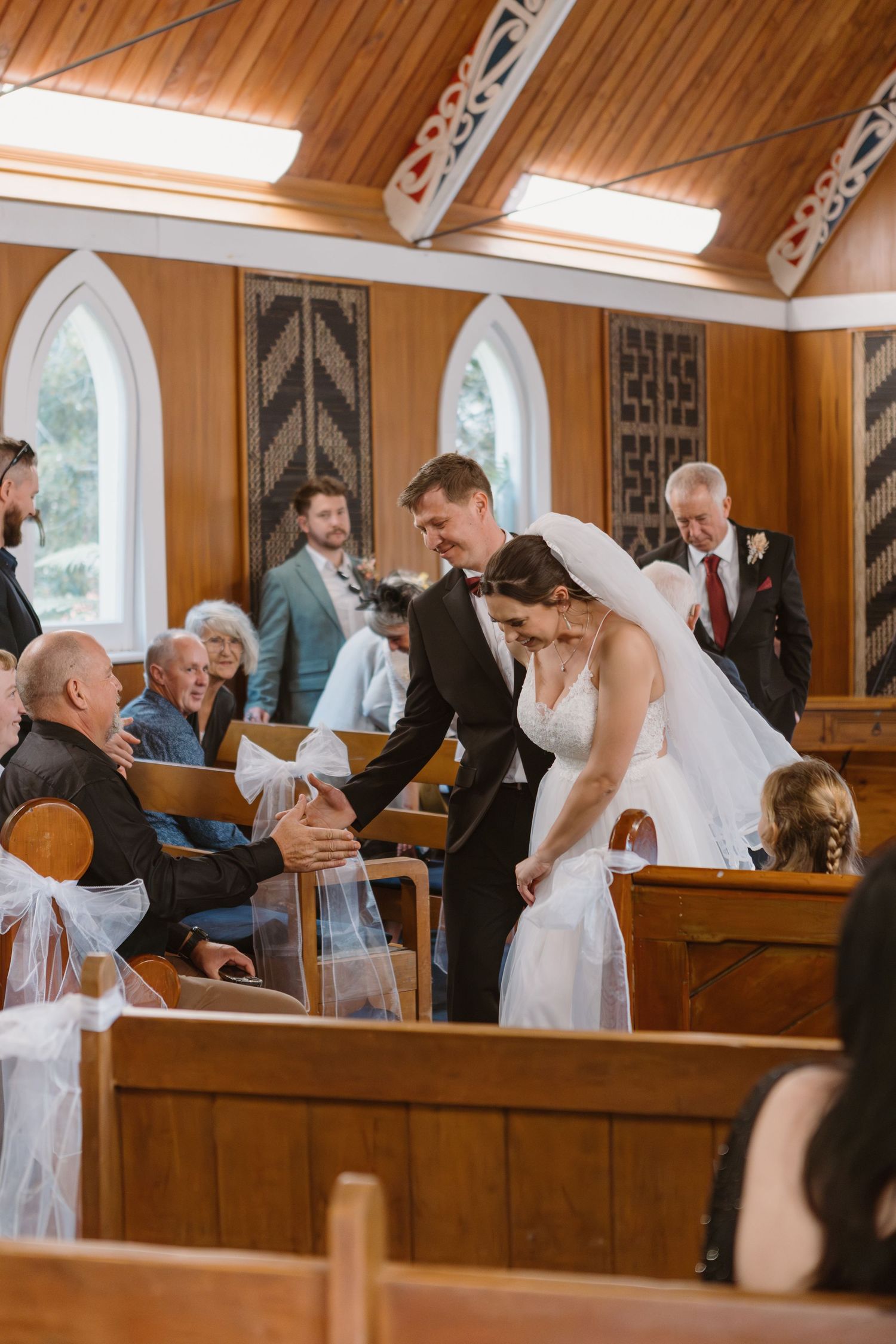 Wedding guests greet the newlyweds in wooden pews inside a church with vaulted ceilings.