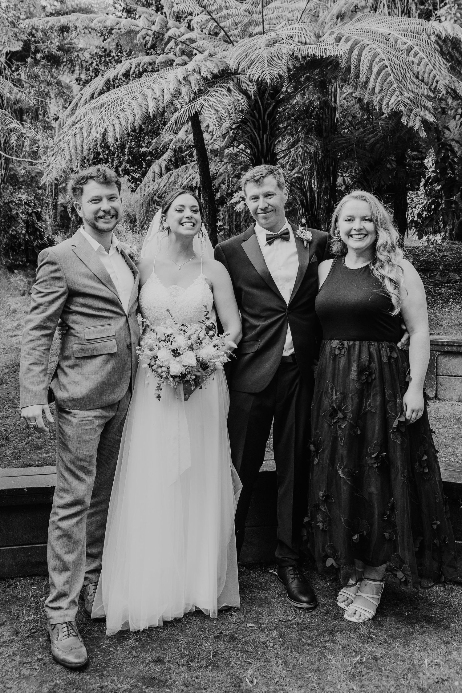 Small wedding party of four poses together in front of tropical foliage at an outdoor garden venue.