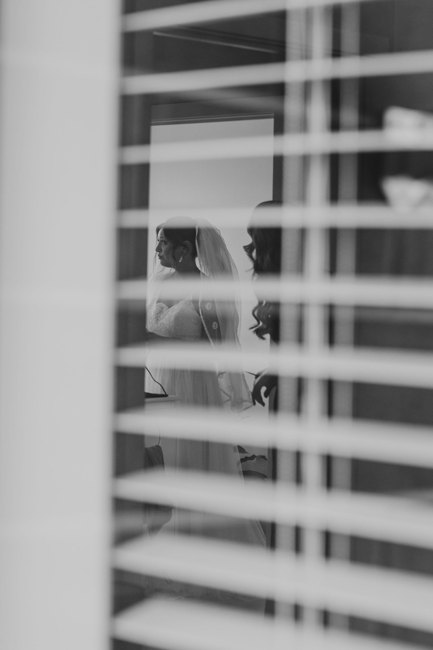 A black and white wedding photo taken through window blinds creating dramatic lines and shadows.