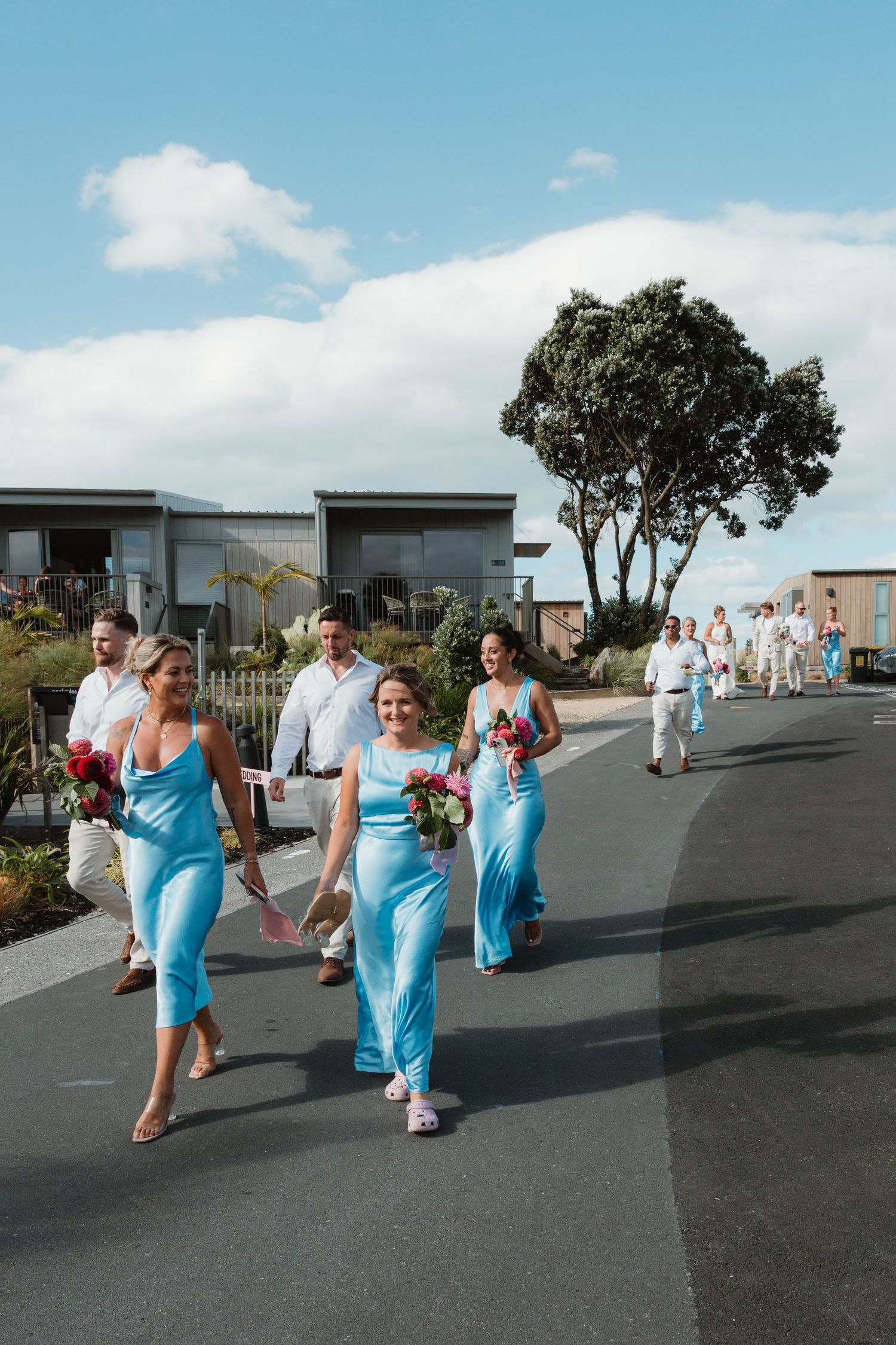 Bridesmaids in blue dresses walk down a road holding bouquets at an outdoor wedding ceremony.
