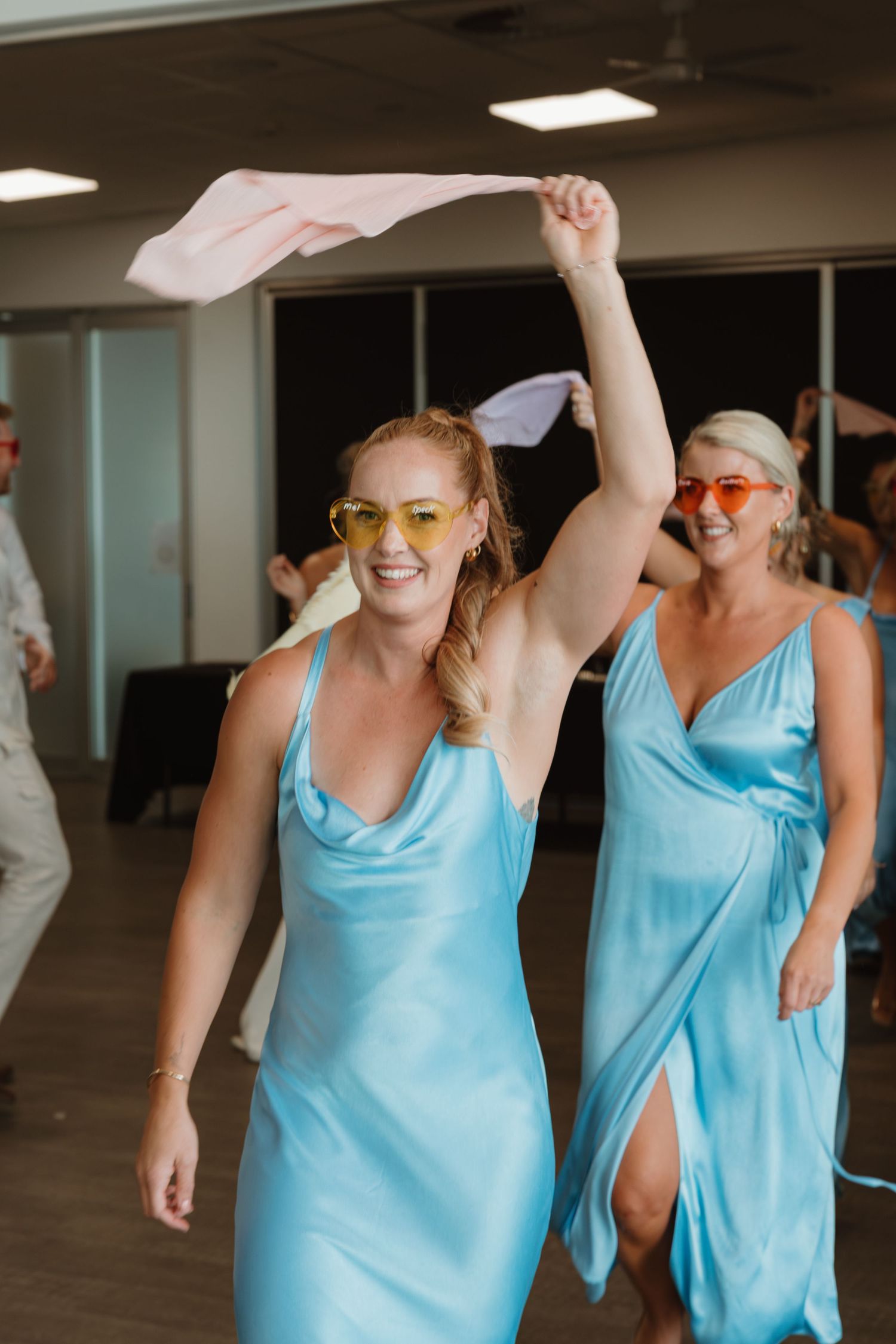 Two bridesmaids in light blue satin dresses and yellow sunglasses dancing joyfully at a wedding reception.