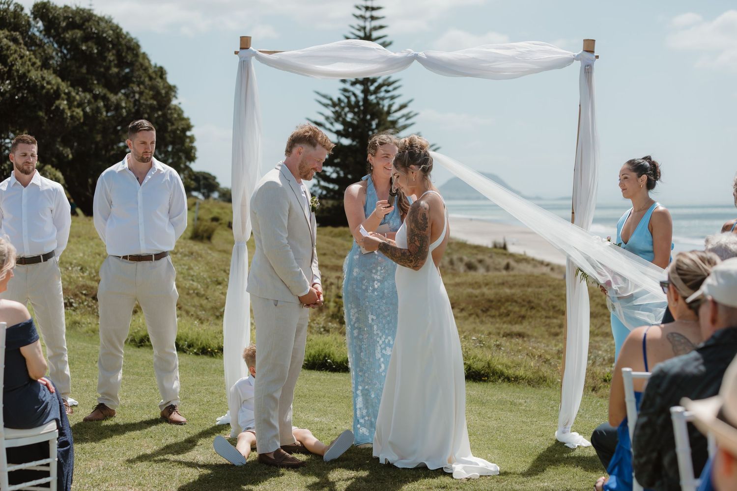 Outdoor wedding ceremony on green lawn with ocean backdrop, couple exchanging vows under white draped arch.