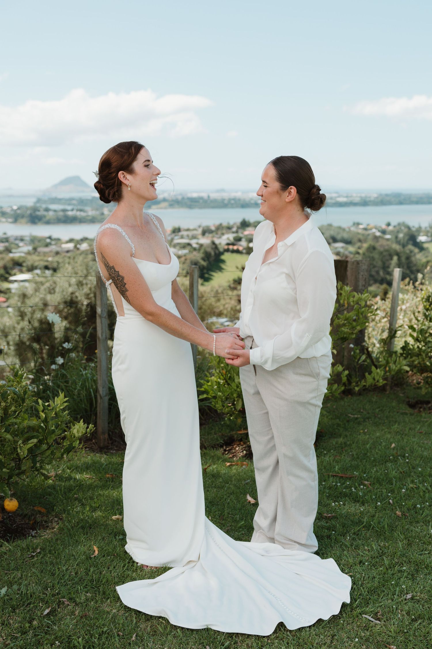 Two brides hold hands outdoors on their wedding day, one in a white gown and one in a cream suit, with a scenic waterfront view.