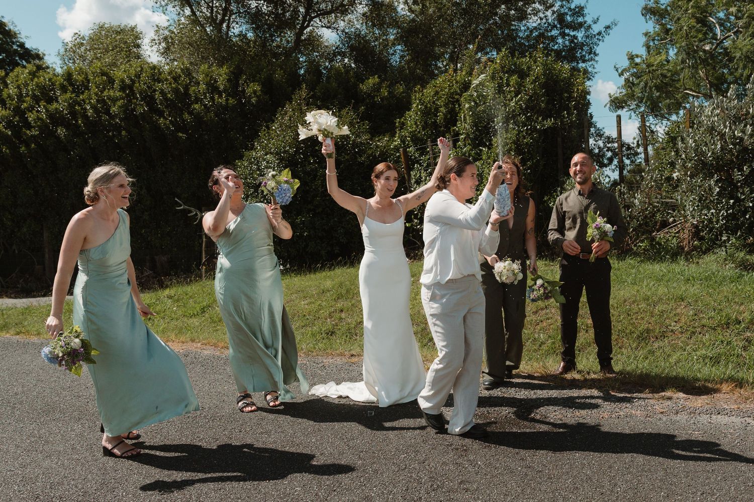 Wedding party celebrating on a road, bride in white gown raising bouquet with bridesmaids in sage green dresses.