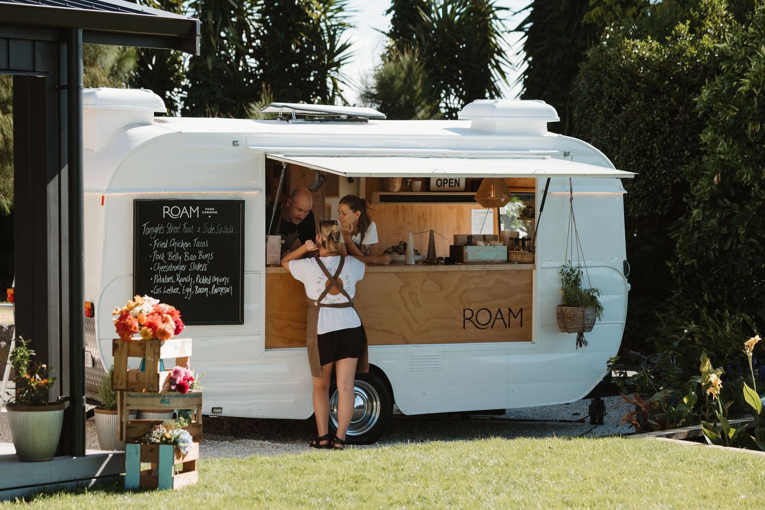 White ROAM food trailer with wooden counter serving customers outdoors in a lush green garden setting.