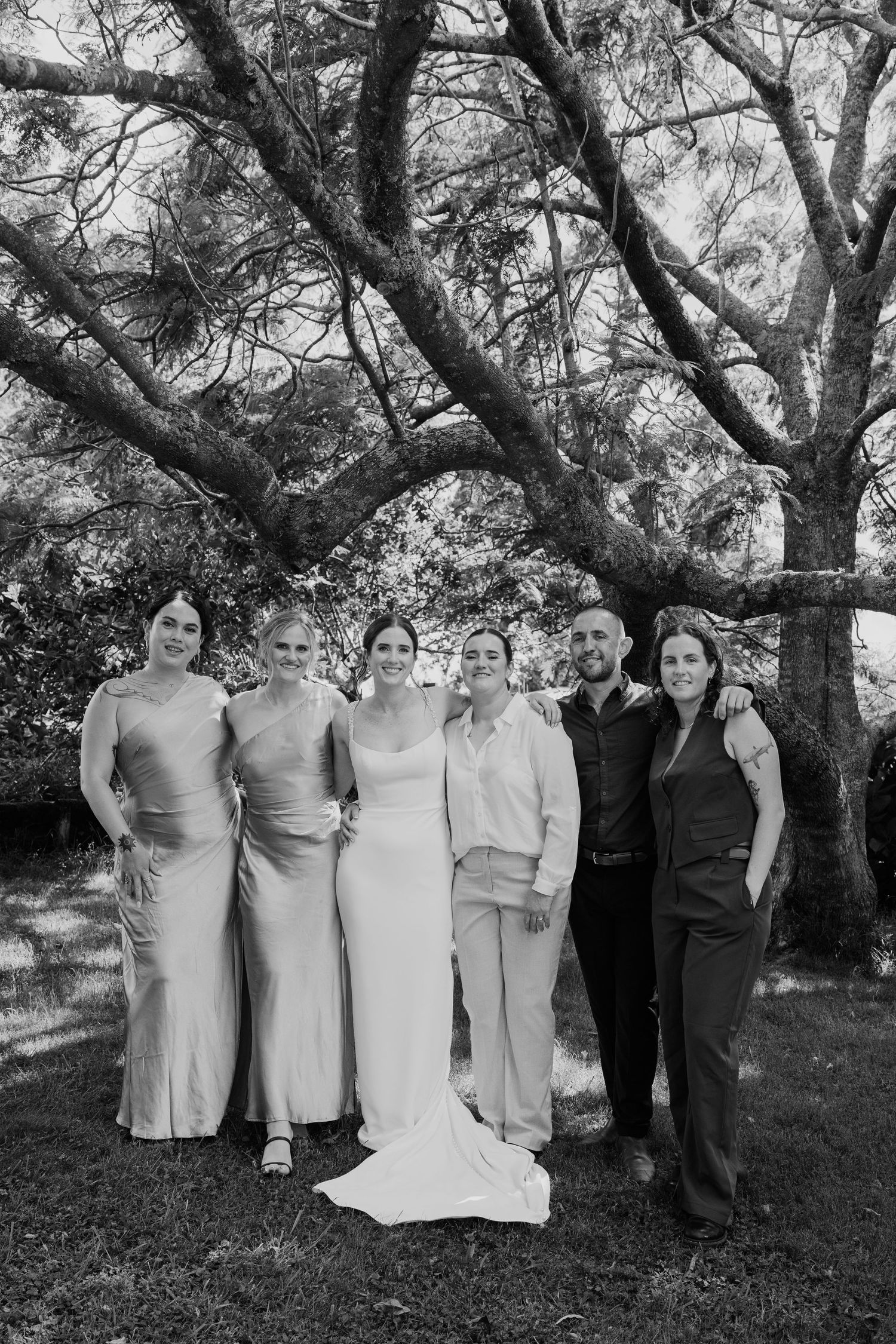 Black and white wedding photo of bride, groom, and bridesmaids posing under a large tree outdoors.