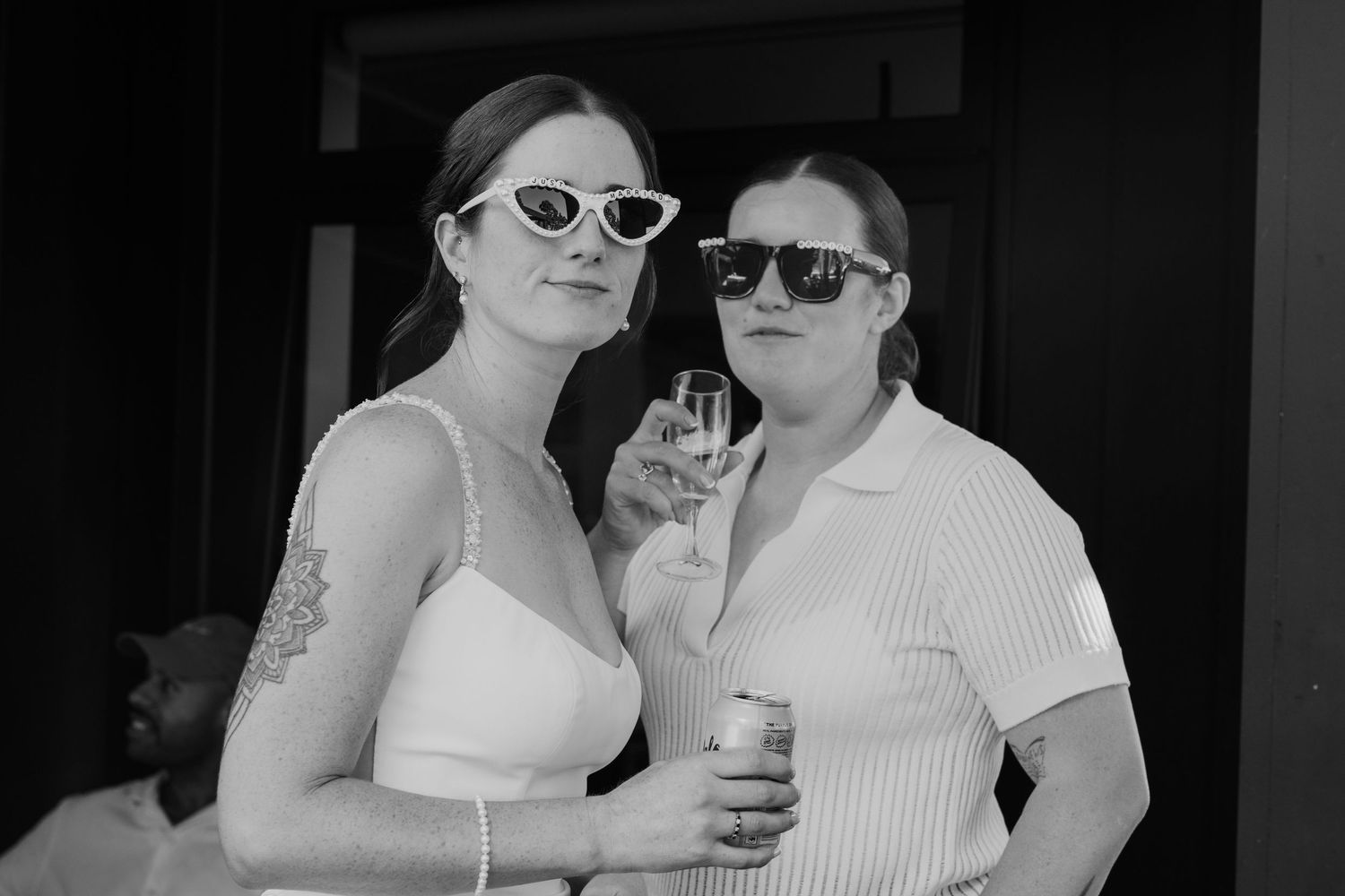 Two women in sunglasses pose together at an outdoor event, one holding a champagne glass, in black and white.